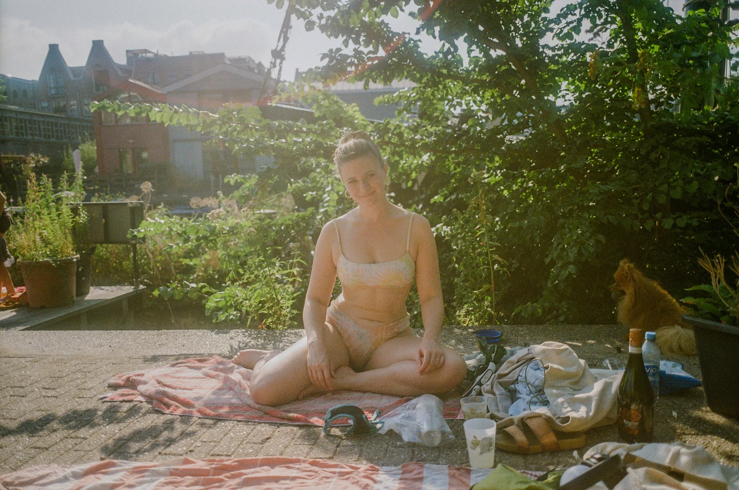 Une femme assise en tailleur sur une serviette, portant un bikini, dans un jardin en plein soleil avec un lionceau en arrière-plan.