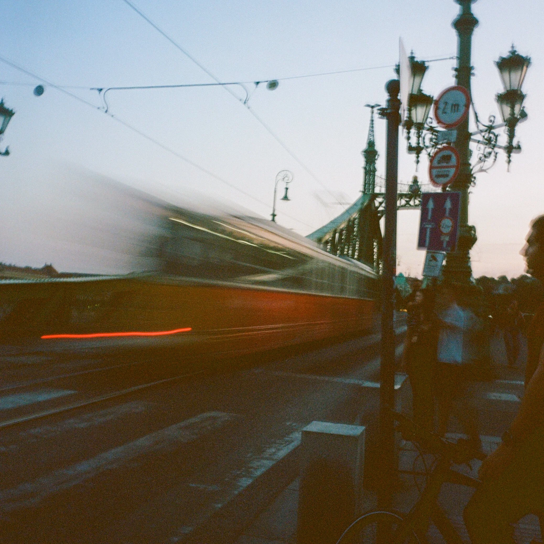 Une photo prise au crépuscule montrant un tramway en mouvement à côté d'un pont, avec des lampadaires et des panneaux de signalisation. Il y a des personnes sur le trottoir, dont un homme à vélo.