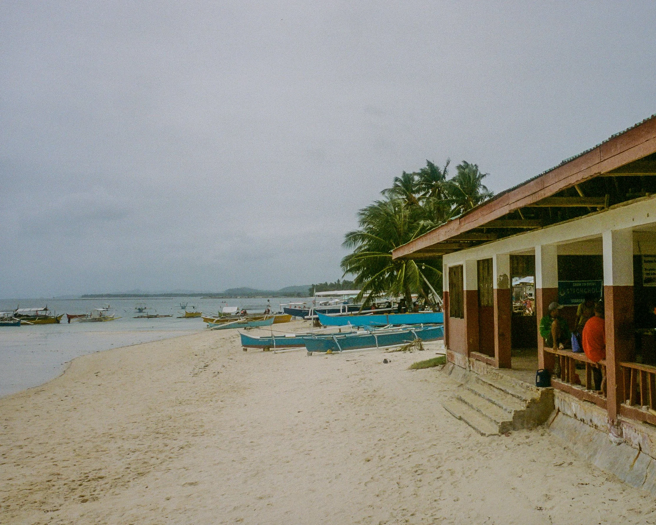  Plage avec plusieurs bateaux de pêche, palmier, bâtiment en bois et personne assise à l'extérieur