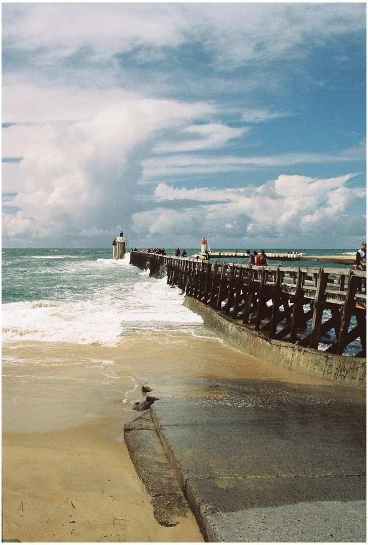 Jetée en bois s'étendant dans la mer avec des personnes assises dessus, deux phares en pierre aux extrémités de la jetée, ciel nuageux, plage de sable en premier plan.