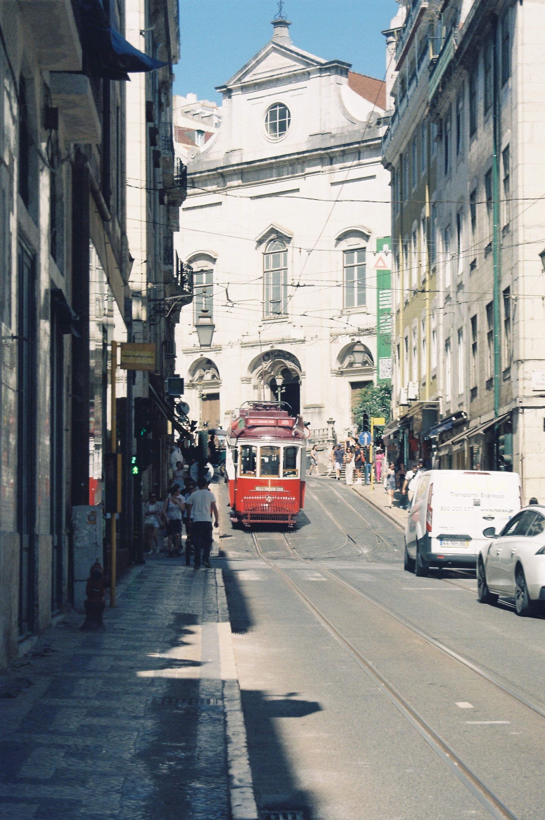 Une rue pavée avec un tramway rouge en montée, entourée de bâtiments vieux avec plusieurs passants et voitures.