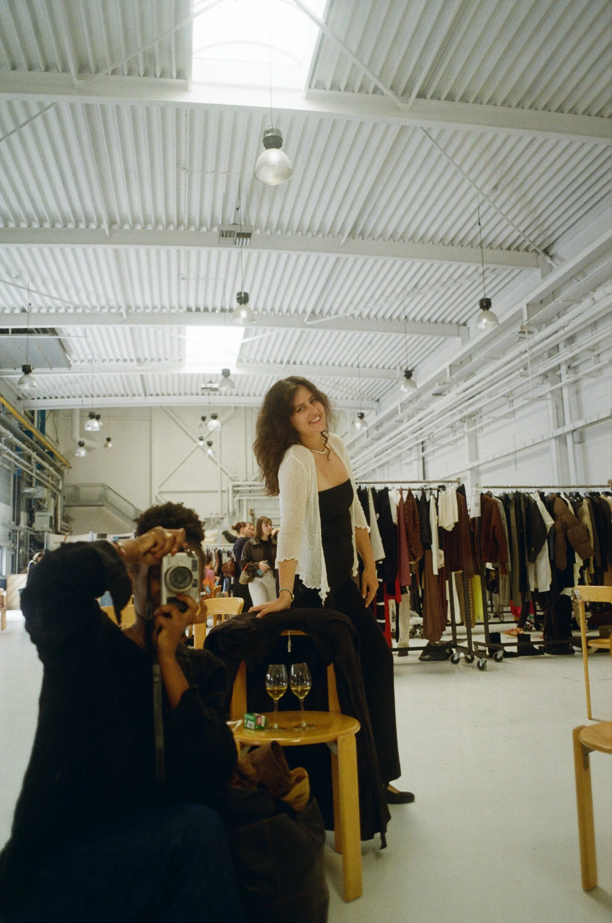 Une femme souriante pose debout avec ses mains sur une chaise dans un grand espace ressemblant à une boutique ou un centre commercial, avec des vêtements sur des racks en arrière-plan. Un photographe s'apprête à prendre une photo d'elle, deux verres 