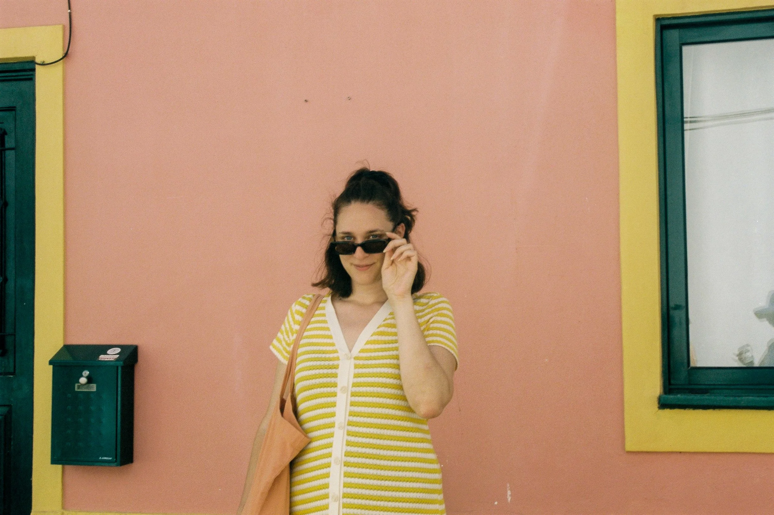 Jeune femme portant une robe rayée jaune et blanc, tenant des lunettes de soleil noires et un sac beige, se tenant devant un mur rose avec des fenêtres vertes.