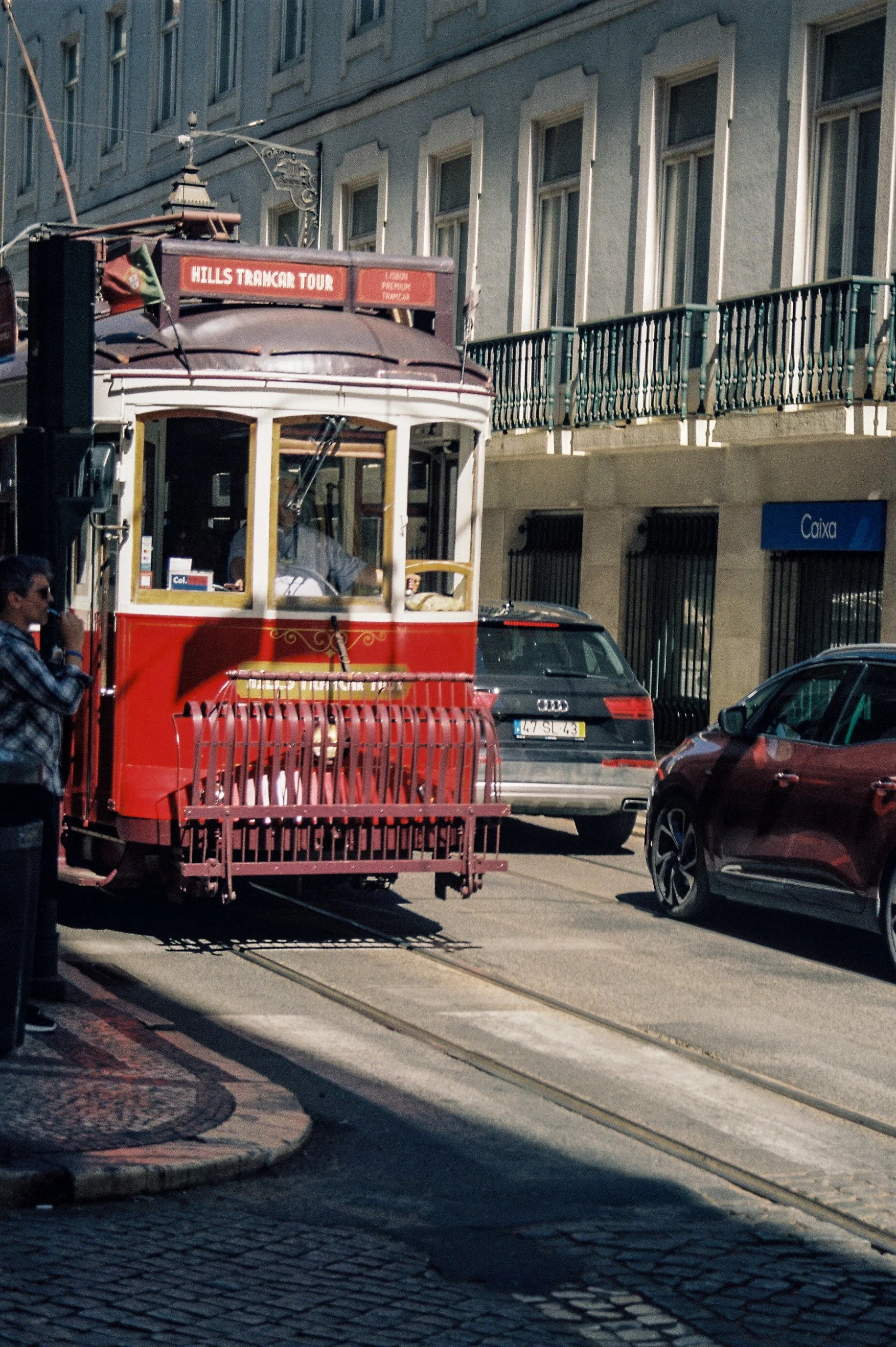 Un tram rouge en circulation dans une rue urbaine avec des voitures garées sur le côté, un homme en vêtements décontractés parlant surement au téléphone ou à une personne, et un bâtiment avec une balustrade en fer forgé. Le tram affiche la destinatio