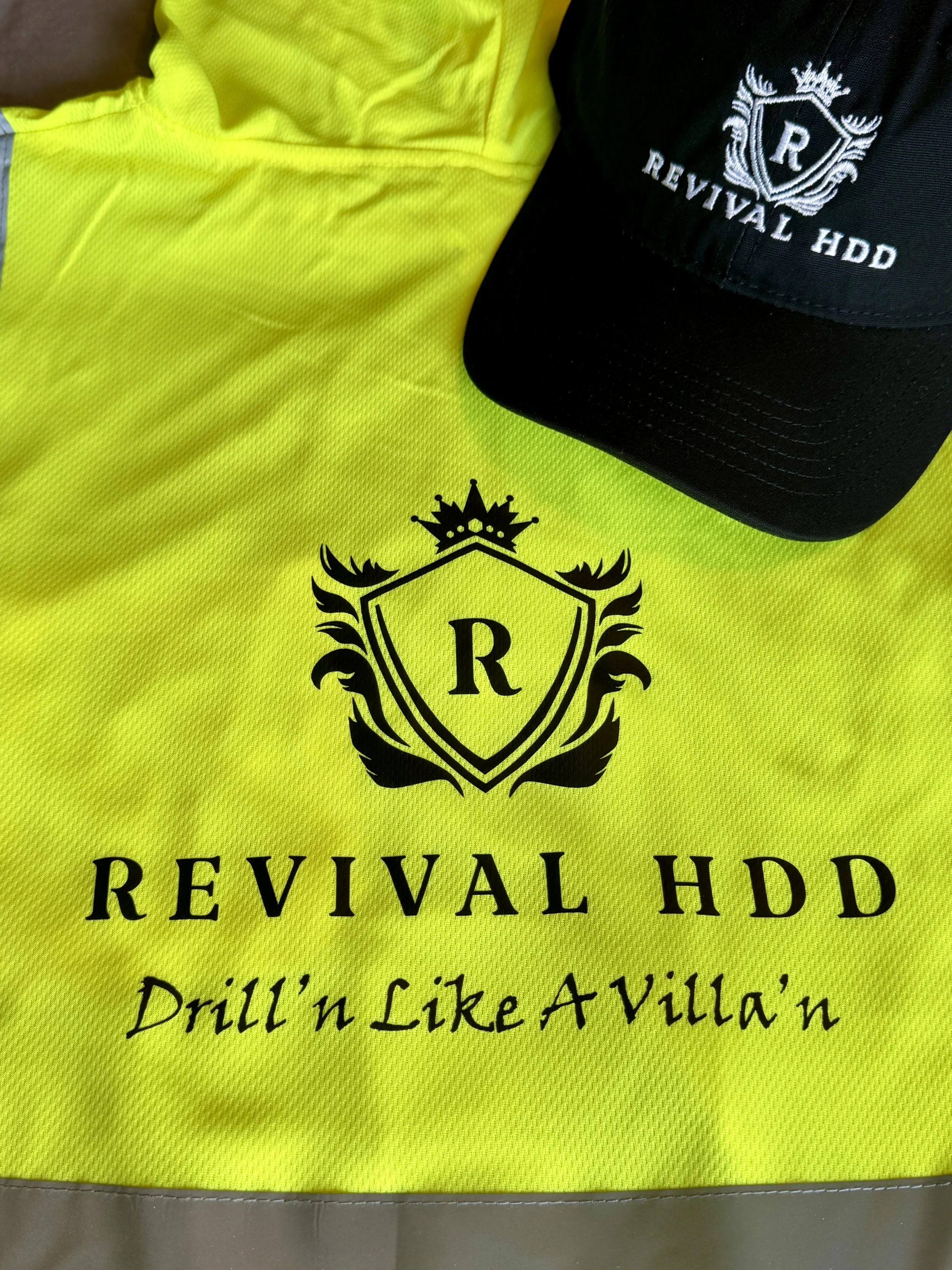 A yellow reflective vest with black text and a logo, and a black cap with white embroidery. The vest reads "REVIVAL HDD" and "Drill 'n Like A Villain." The cap also displays the text "REVIVAL HDD" along with a crest and the letter "R."