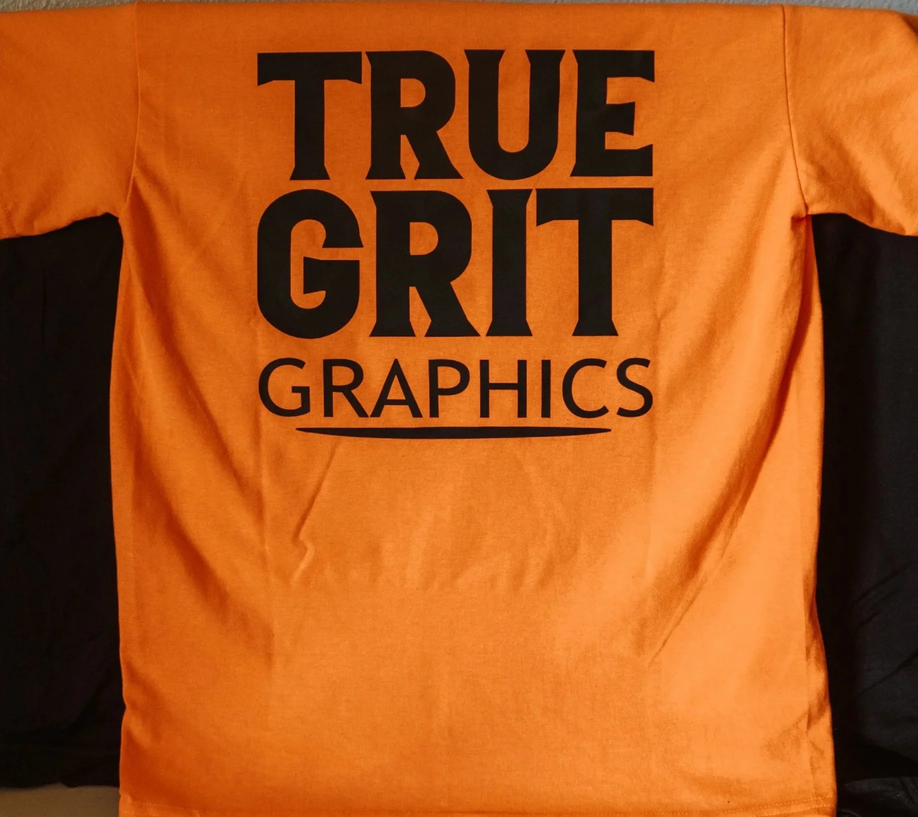 Orange T-shirt with black text that reads 'TRUE GRIT GRAPHICS' on the back