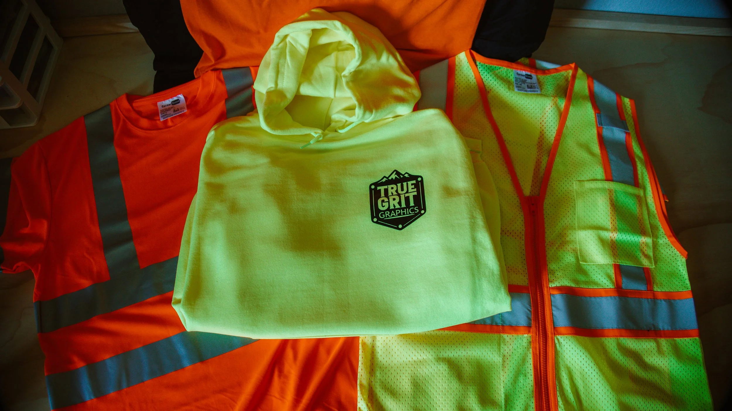 High-visibility safety vest, orange and yellow hoodie with 'True Grit Graphics' logo, and orange and gray safety shirt laid out on a wooden surface.
