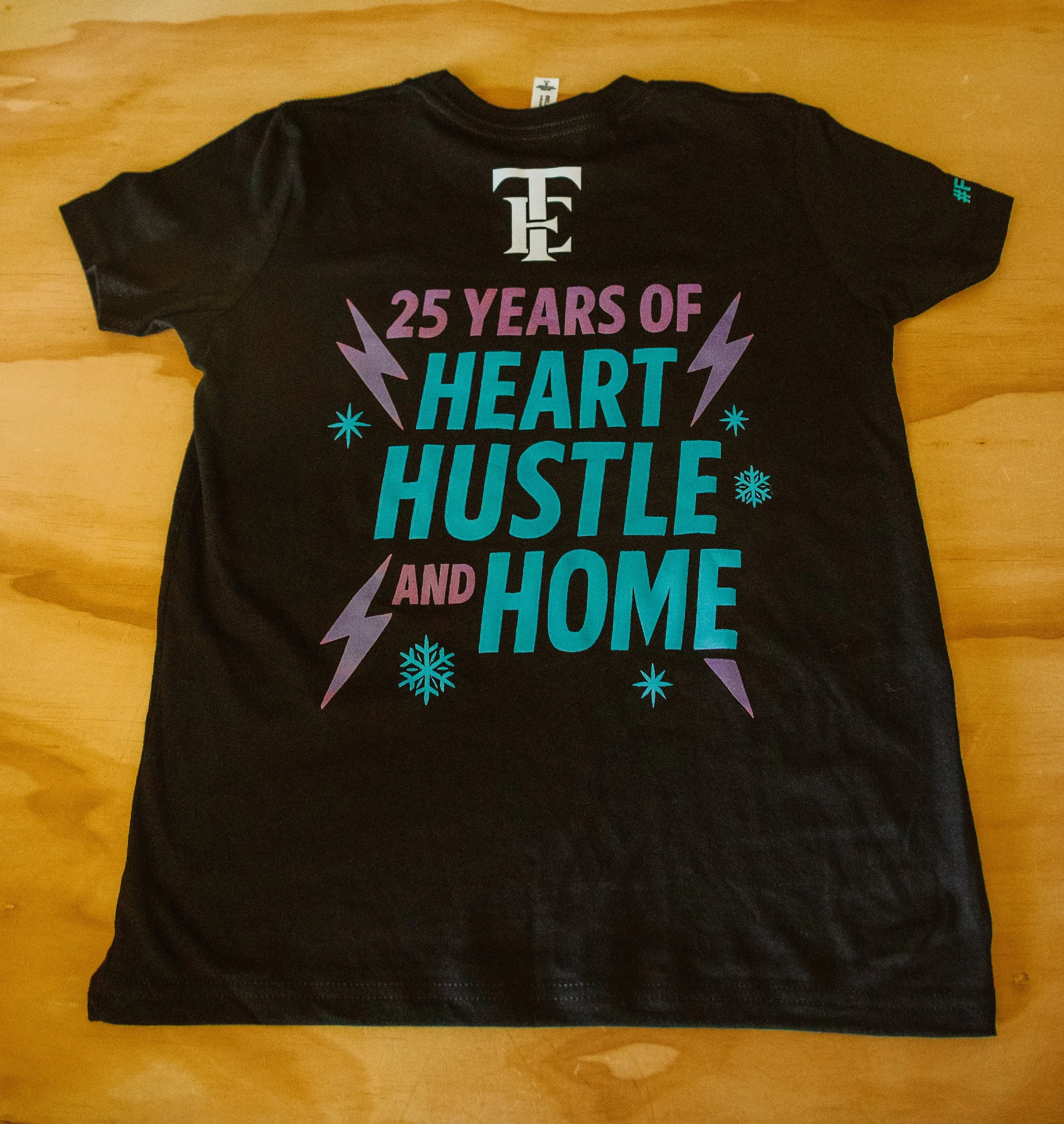 Black T-shirt for a cheerleading gym with text celebrating 25 years of heart, hustle, and home, decorated with purple lightning bolts, snowflakes, and stars.