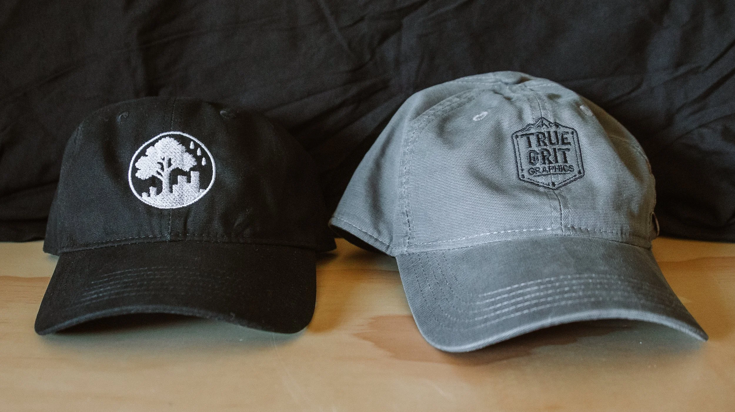 Two baseball caps on a wooden surface, one black with a white tree logo and the other gray with a 'True Grit Graphics' patch.