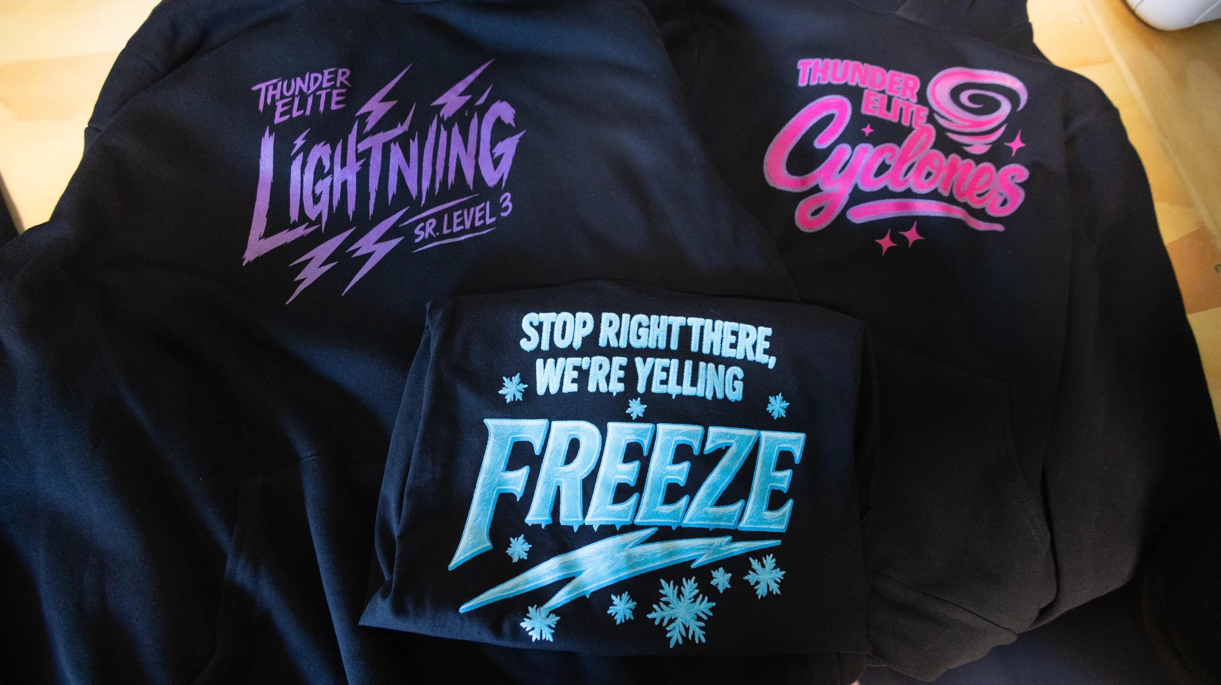 Three black hoodies with colorful designs and text. One hoodie reads 'Thunder Elite Lightning Sr. Level 3' in purple with lightning bolt graphics. The second says 'Thunder Elite Cyclones' in pink with a swirling graphic. The third states 'Stop right
