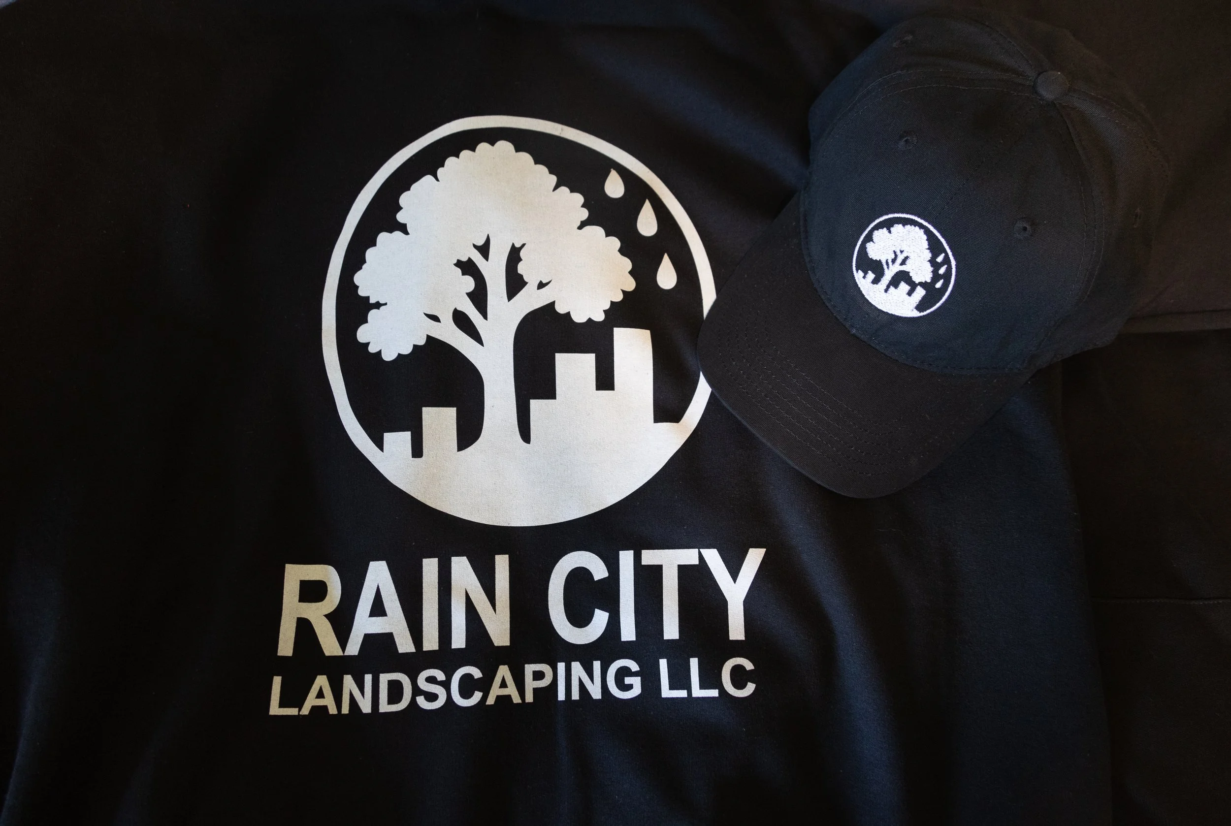 A black T-shirt and a black baseball cap both display a logo of a tree and cityscape with raindrops, and the text "RAIN CITY LANDSCAPING LLC" on the shirt.
