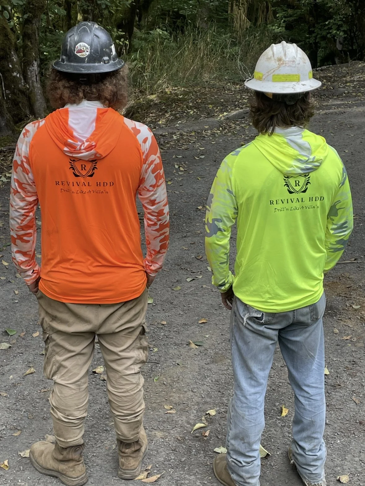 Two construction workers standing outdoors on a dirt path, viewed from behind. They are wearing hard hats and bright-colored hooded jackets with the logo 'Revival HDD' and the slogan 'Drill'n Like a Villain,' with camouflage pattern on the sleeves. T