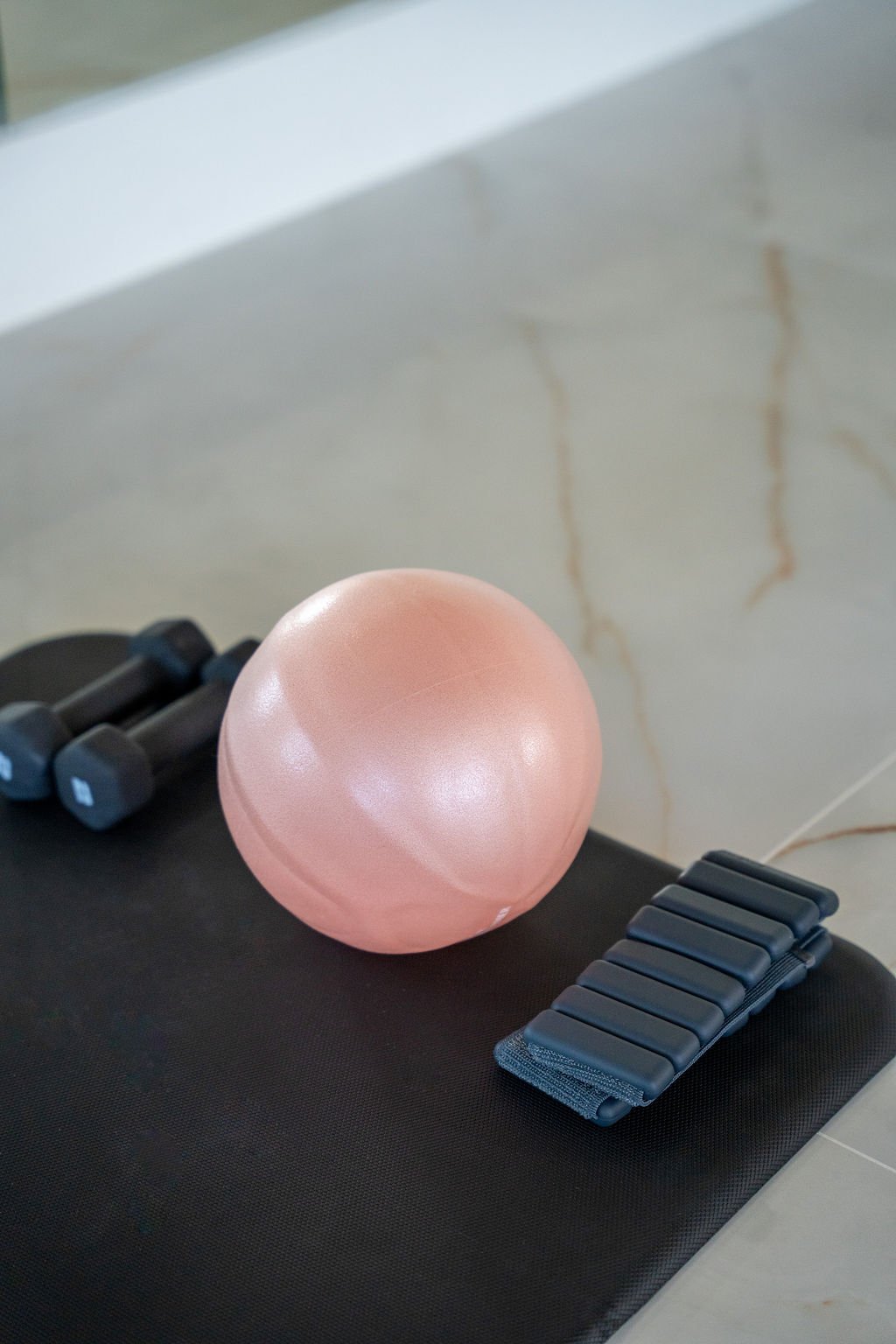 Pink exercise ball, two black dumbbells, and a black resistance band on a black workout mat.