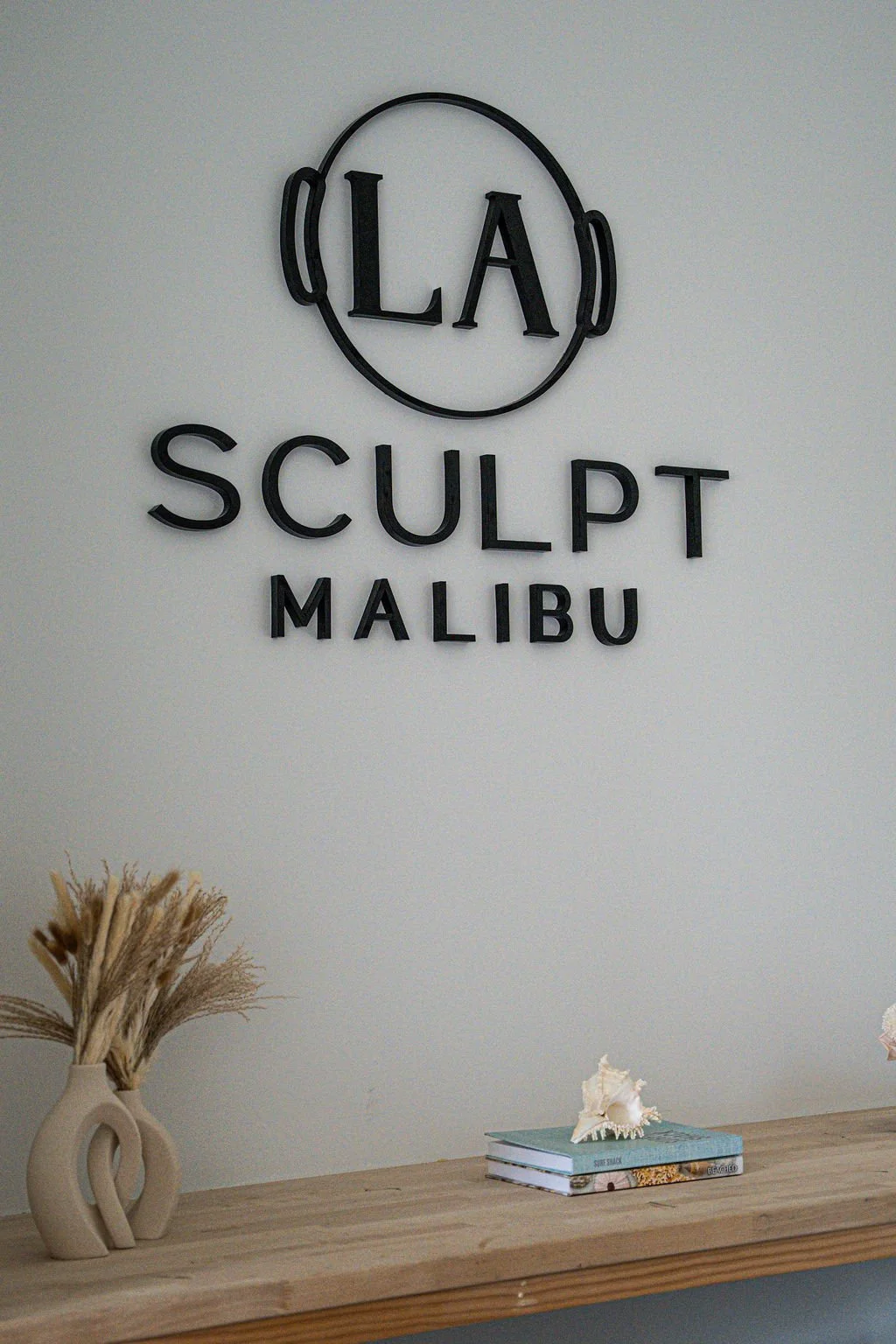 Decorative sign on a wall with text 'LA SCULPT MALIBU' and a logo of a pilates ring. A wooden shelf below holds a beige vase with dried grass and a small stack of books with a shell on top.