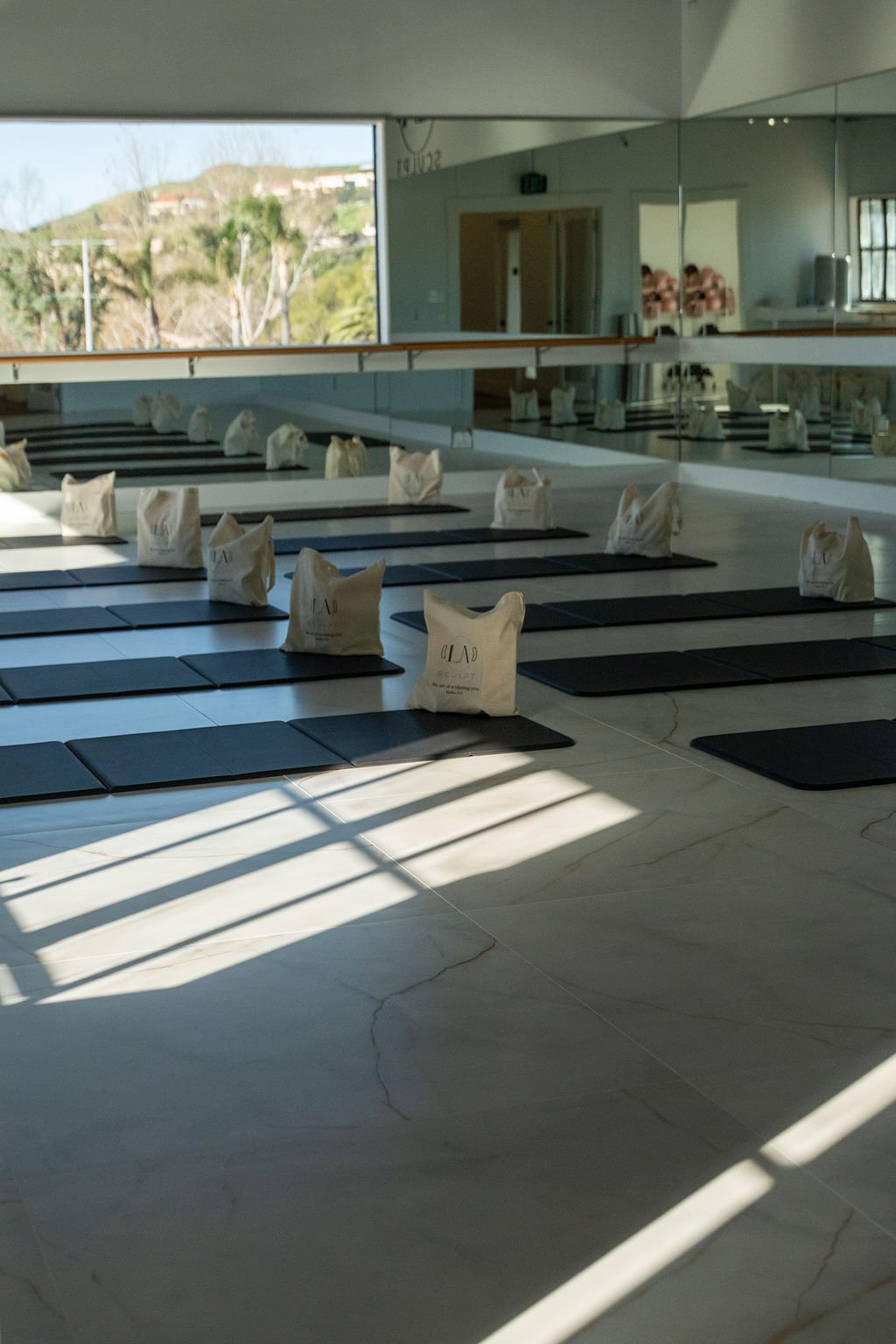 Empty pilates studio with black mats and branded bags on each mat, sunlight streaming through windows, and mirrored wall.