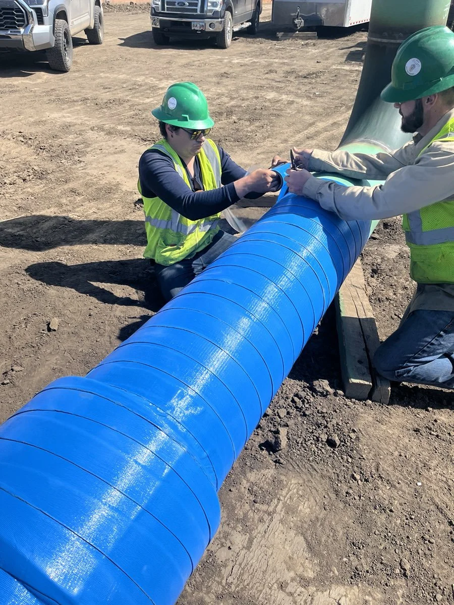 Two construction workers wearing green hard hats and safety vests are installing a large blue pipe on the ground outdoors, with vehicles parked in the background.