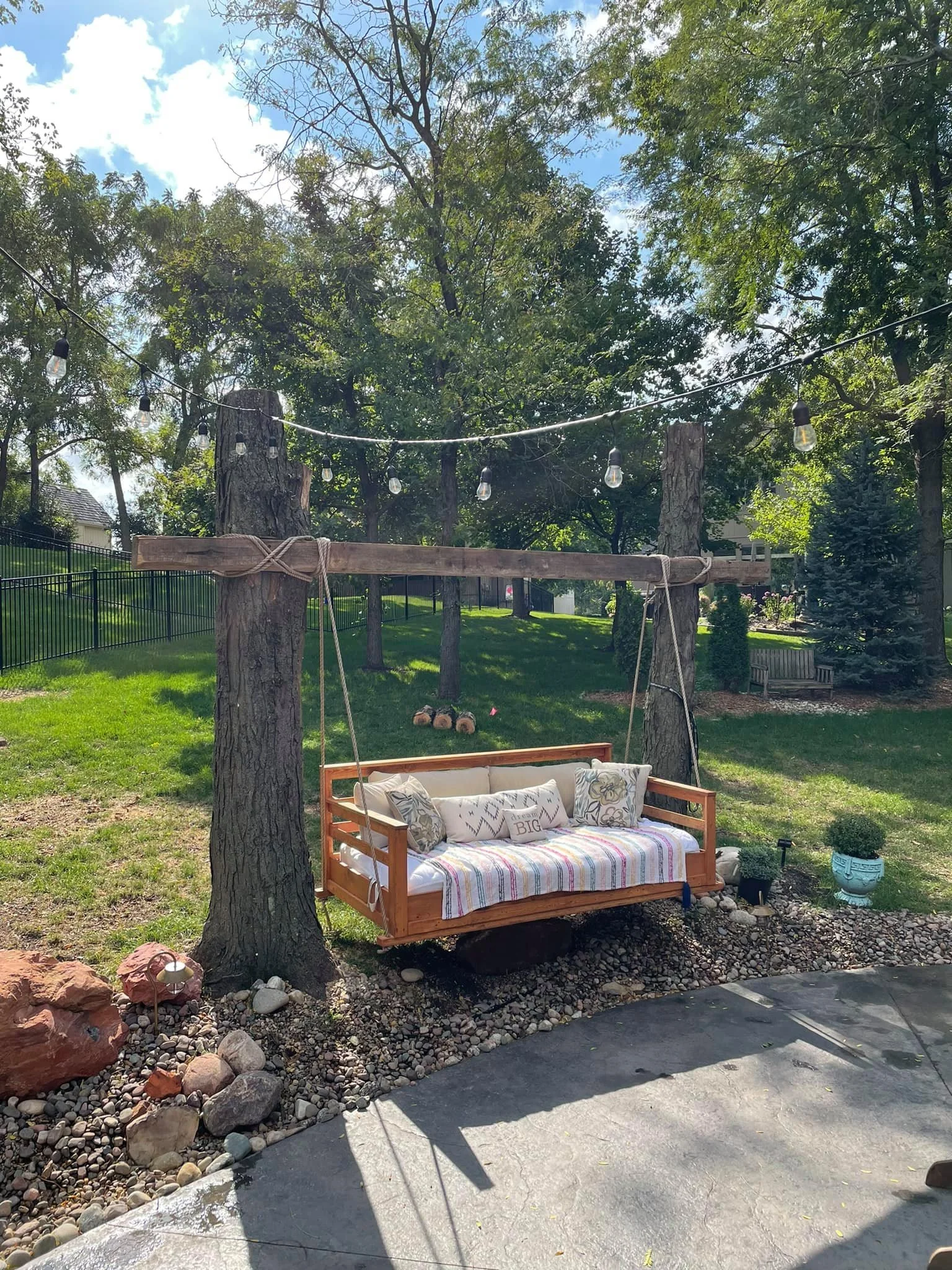 Outdoor backyard scene with a wooden swing hanging from a wooden beam supported by two trees, string lights above, a grassy yard with trees, a bench, and potted plants.