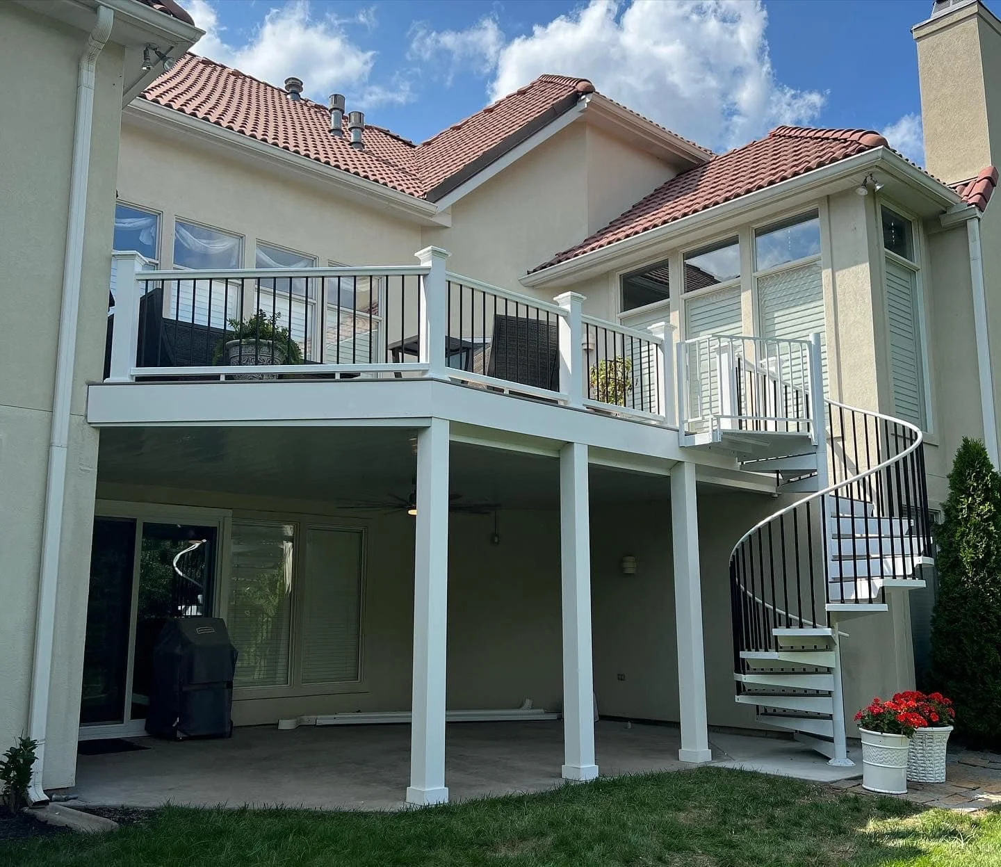 Back view of a two-story house with a white deck and black railing, spiral staircase, outdoor furniture, and a potted plant with red flowers.