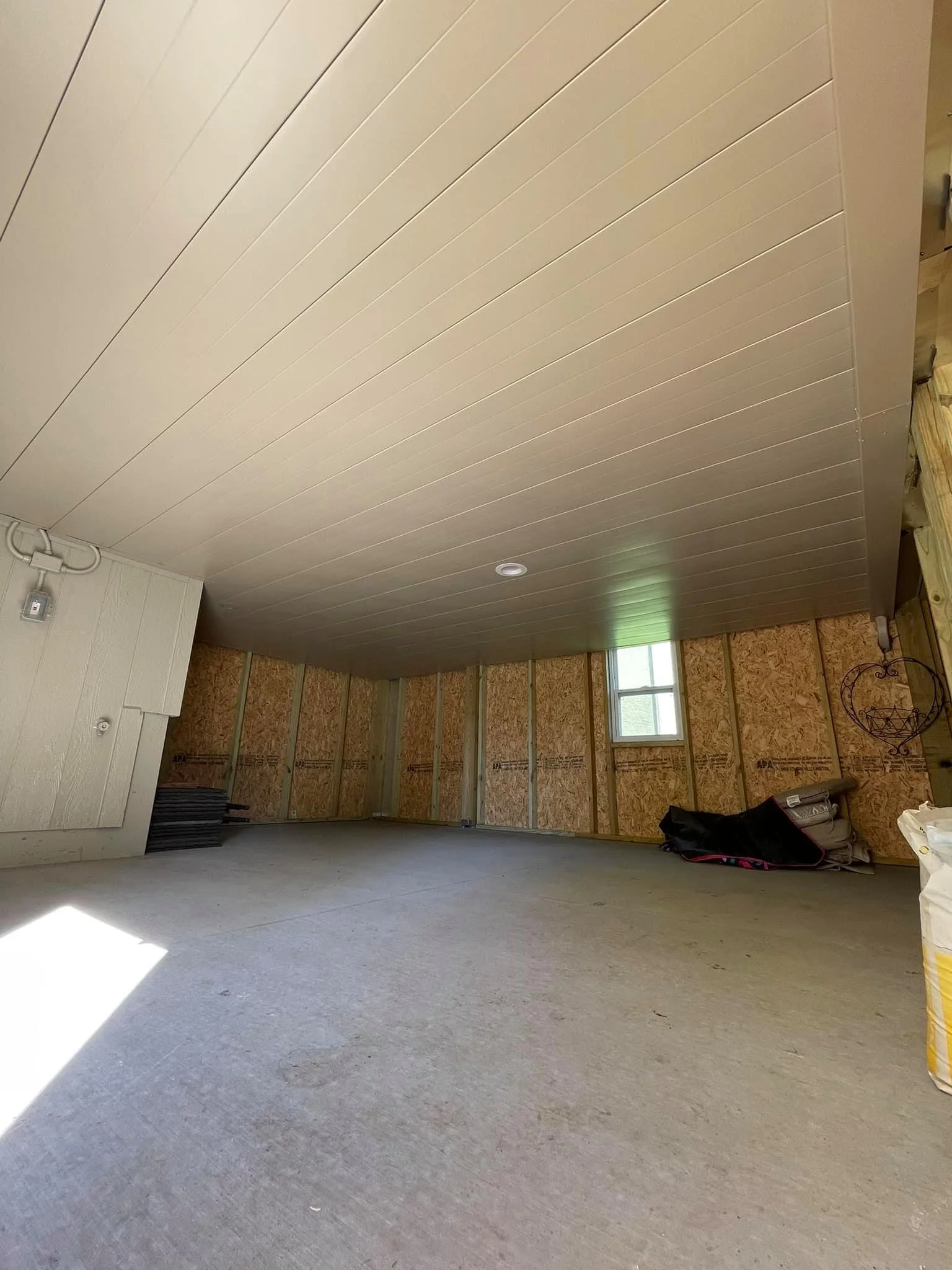 An unfinished interior room with a tan ceiling, exposed wall studs, a small window, and construction materials on the floor.