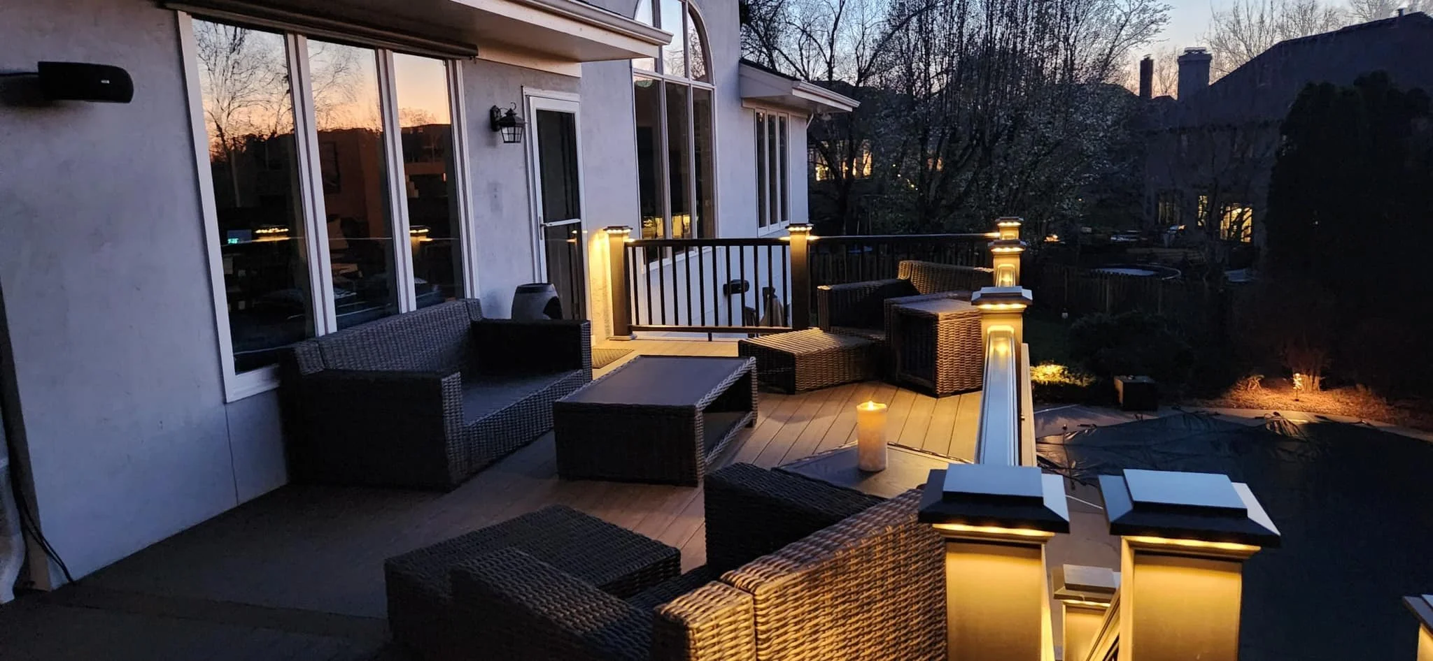 Outdoor patio at dusk with wicker seating, a candle, and ambient lighting on posts, overlooking a backyard with a pool cover and trees.