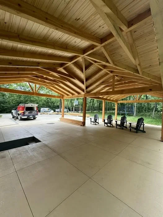 Open-air wooden pavilion with a high vaulted ceiling, concrete flooring, and several black Adirondack chairs. There is a parking lot with a truck and cars outside, surrounded by trees.