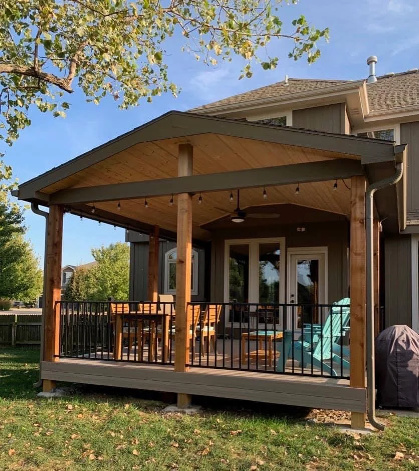 A wooden backyard patio with a covered roof, outdoor furniture, a ceiling fan, and string lights, attached to a gray house with sliding glass doors. There is a green lawn with fallen leaves and trees in the background.
