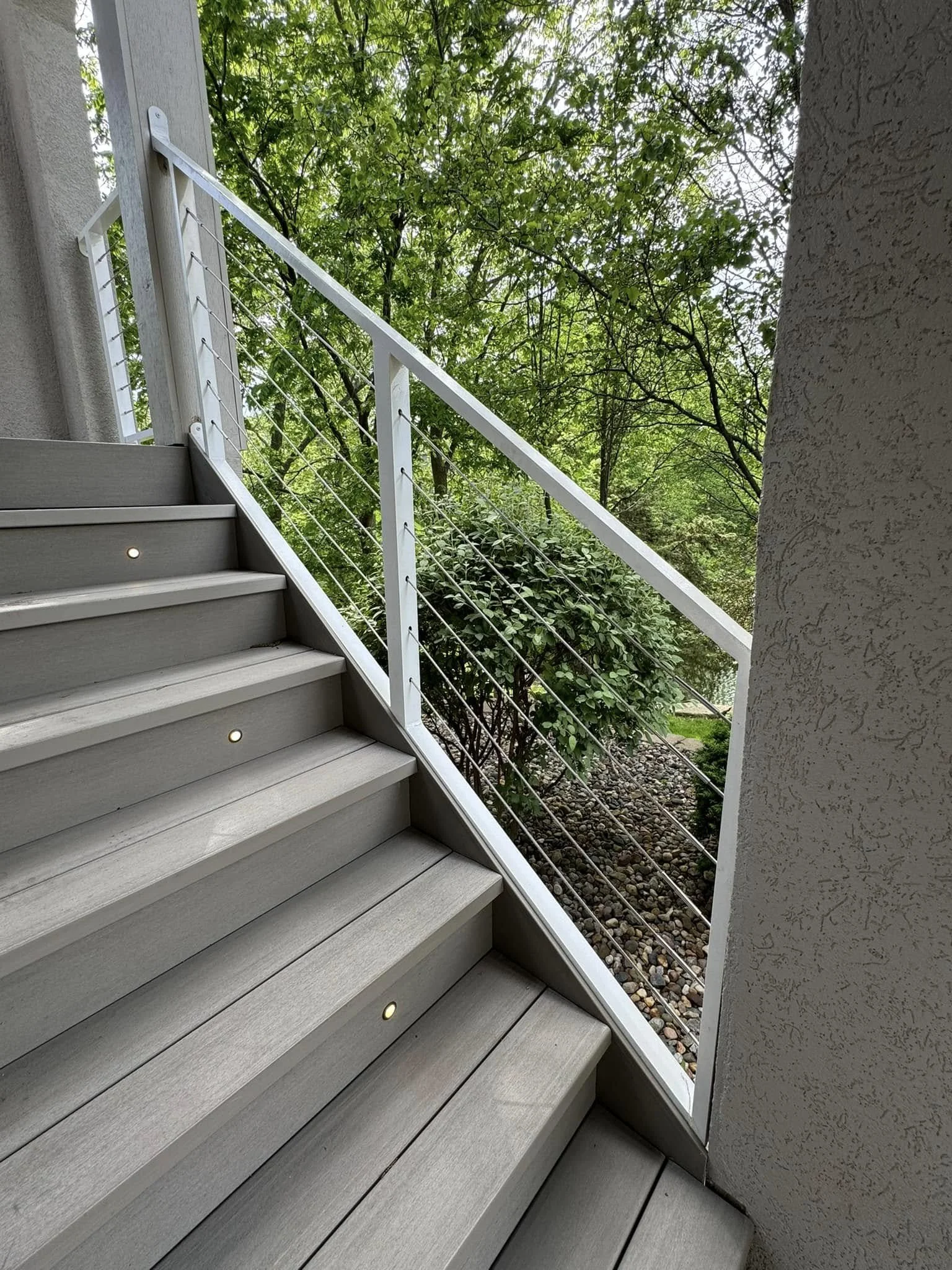 Outdoor staircase with gray wooden steps, metal railing with horizontal wires, and lush green trees in the background.