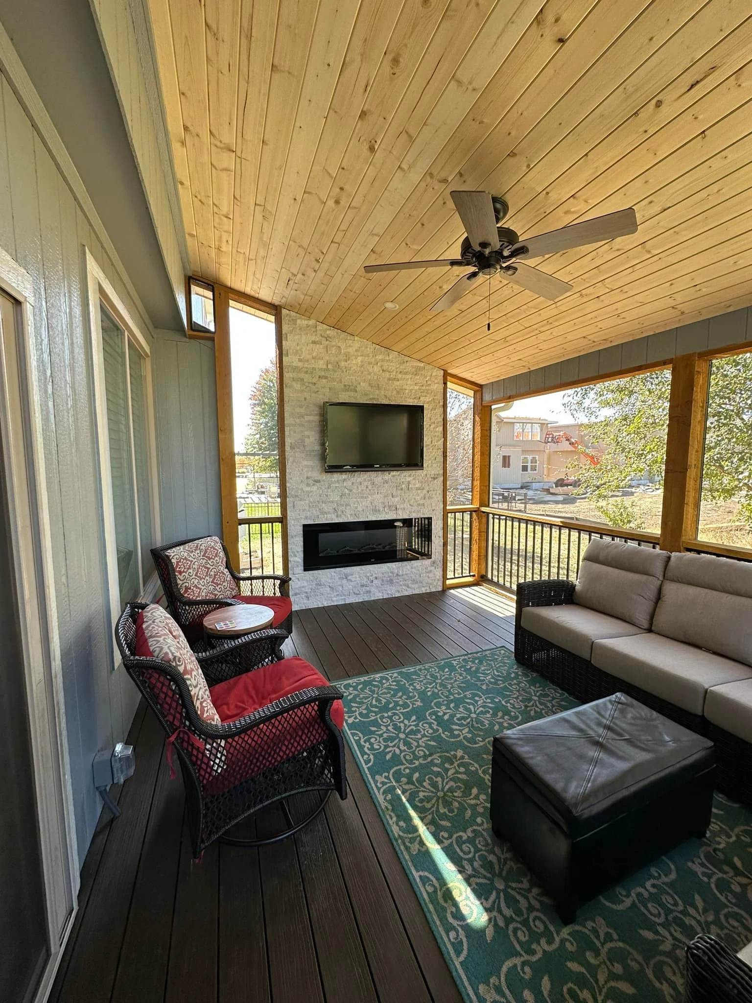 Covered porch with wooden ceiling, ceiling fan, wall-mounted TV, electric fireplace, wicker sofa with beige cushions, two red cushioned wicker chairs with patterned pillows, teal patterned area rug, and railing overlooking a yard with trees and neigh