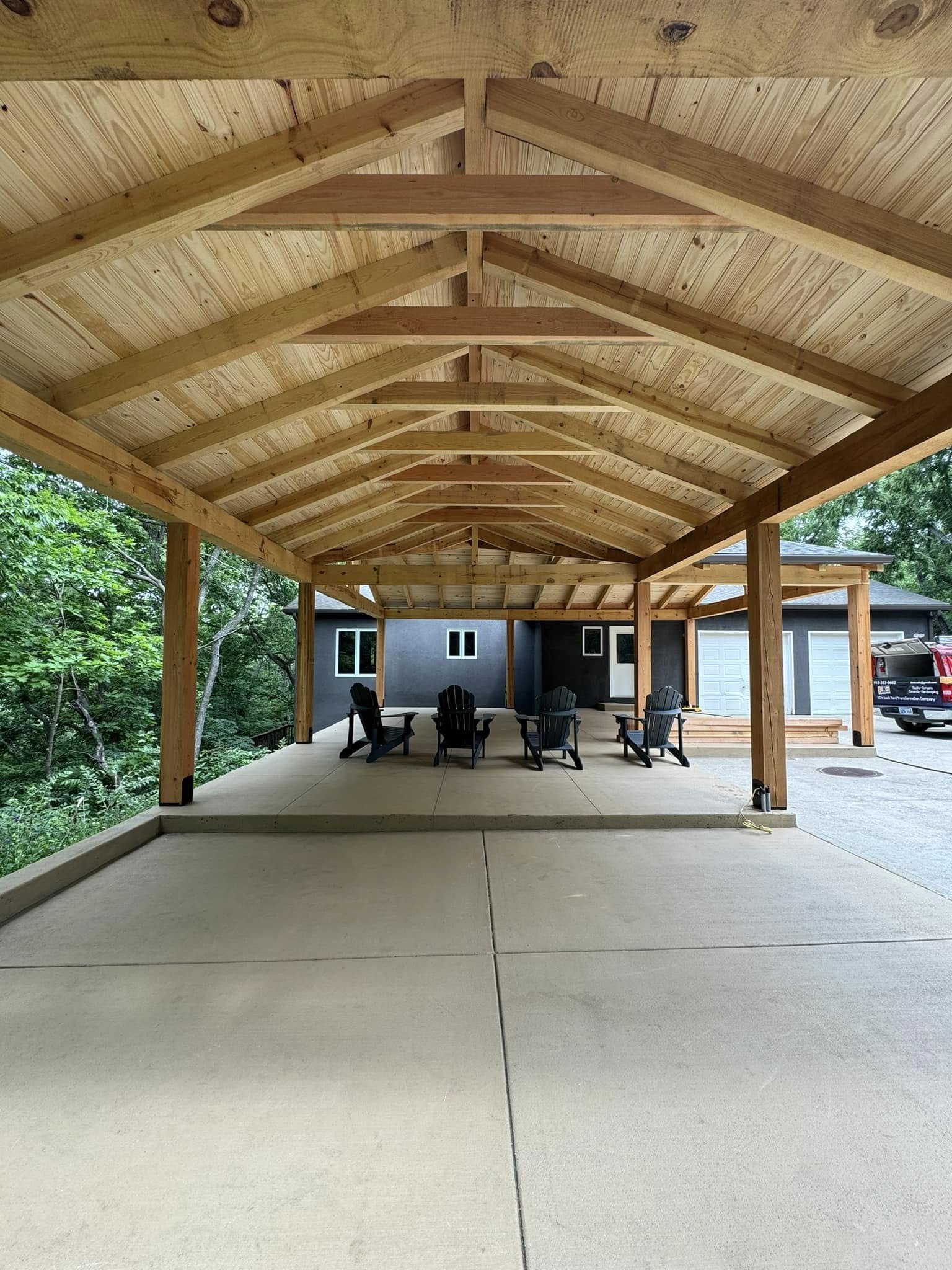Covered patio with wooden beams and a concrete floor, featuring several black chairs arranged around a table, with a house and a vehicle in the background surrounded by trees.