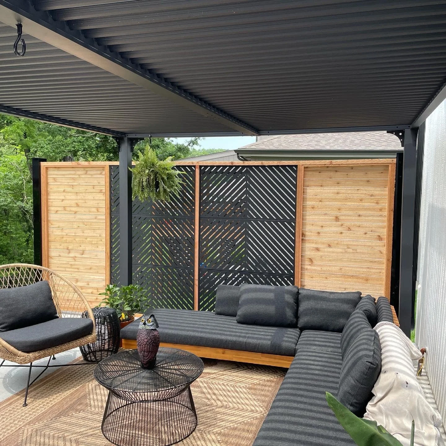 Outdoor patio area with black cushioned sectional sofa, rattan chair with cushion, black wire coffee table with an owl decoration, potted plants, hanging fern, and wooden privacy fences with a black lattice design in a backyard.