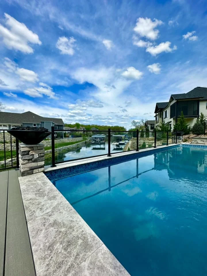 Residential swimming pool with a view of a canal and houses in the background under a blue sky with clouds.