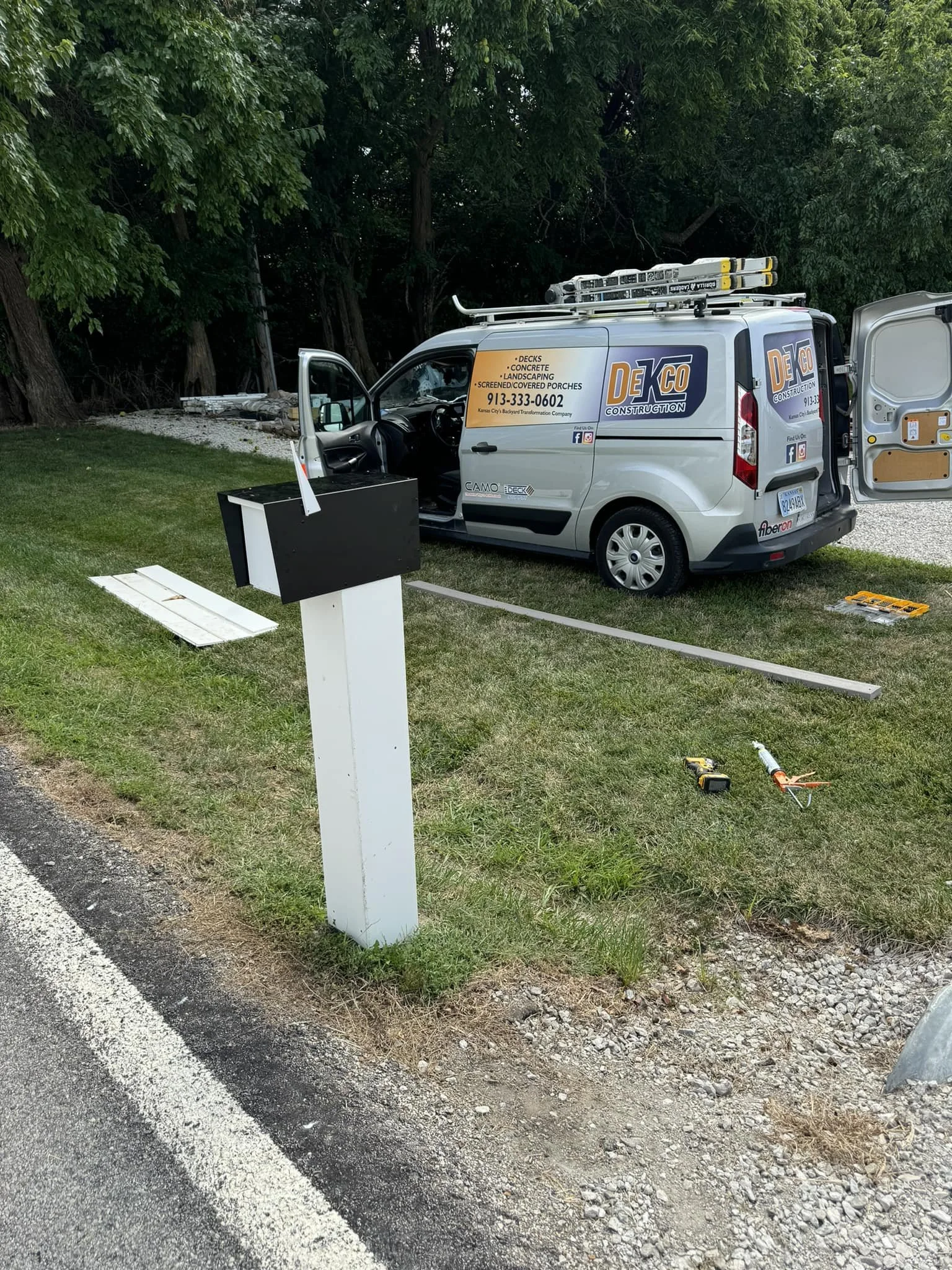 Work van parked on grassy area with open side door, tools and equipment on ground, mailbox nearby, and trees in background.