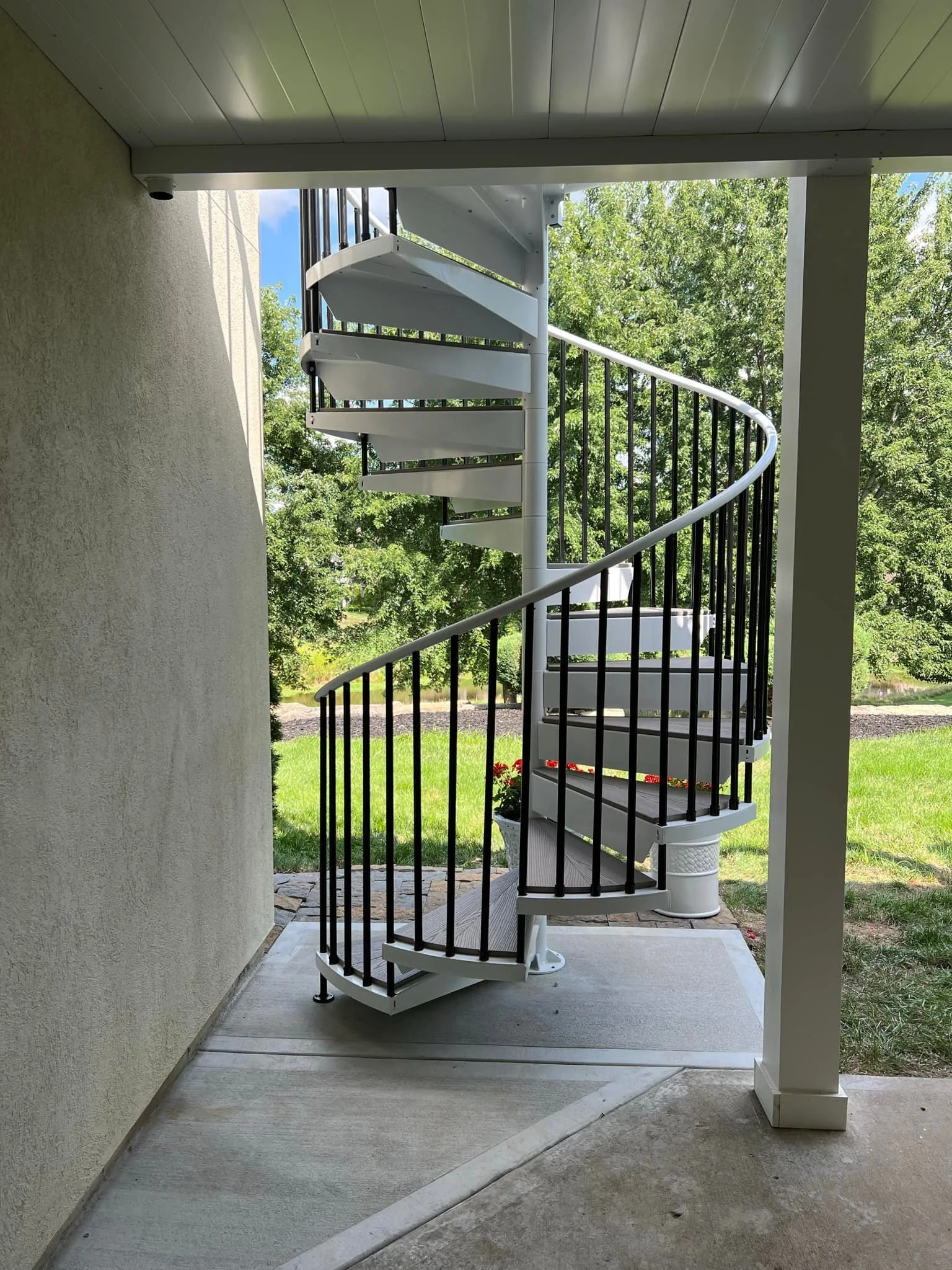 White outdoor spiral staircase with black railing leading to an upper level, viewed from a covered porch area with a concrete floor. Green trees and blue sky are visible in the background.