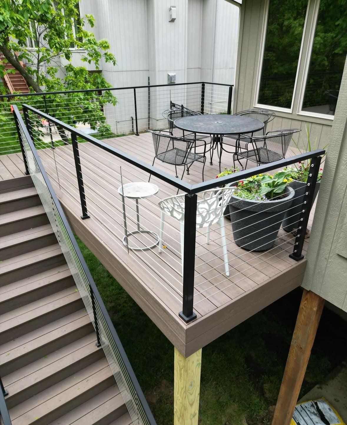 A wooden outdoor deck with black railing, featuring a round table with four chairs, a small white side table, a large black planter with greenery, and a white decorative chair, overlooking green trees and a neighboring building.