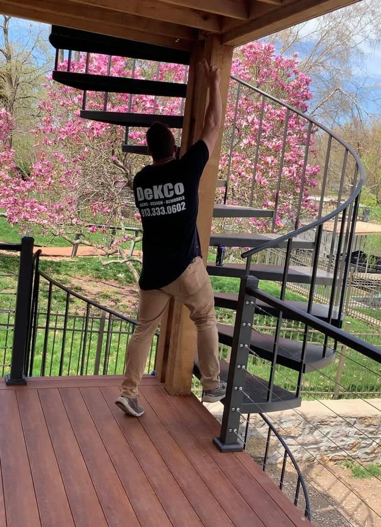 A person with a black shirt and khaki pants working on an outdoor spiral staircase made of metal and wood, surrounded by blooming pink flowers and trees.