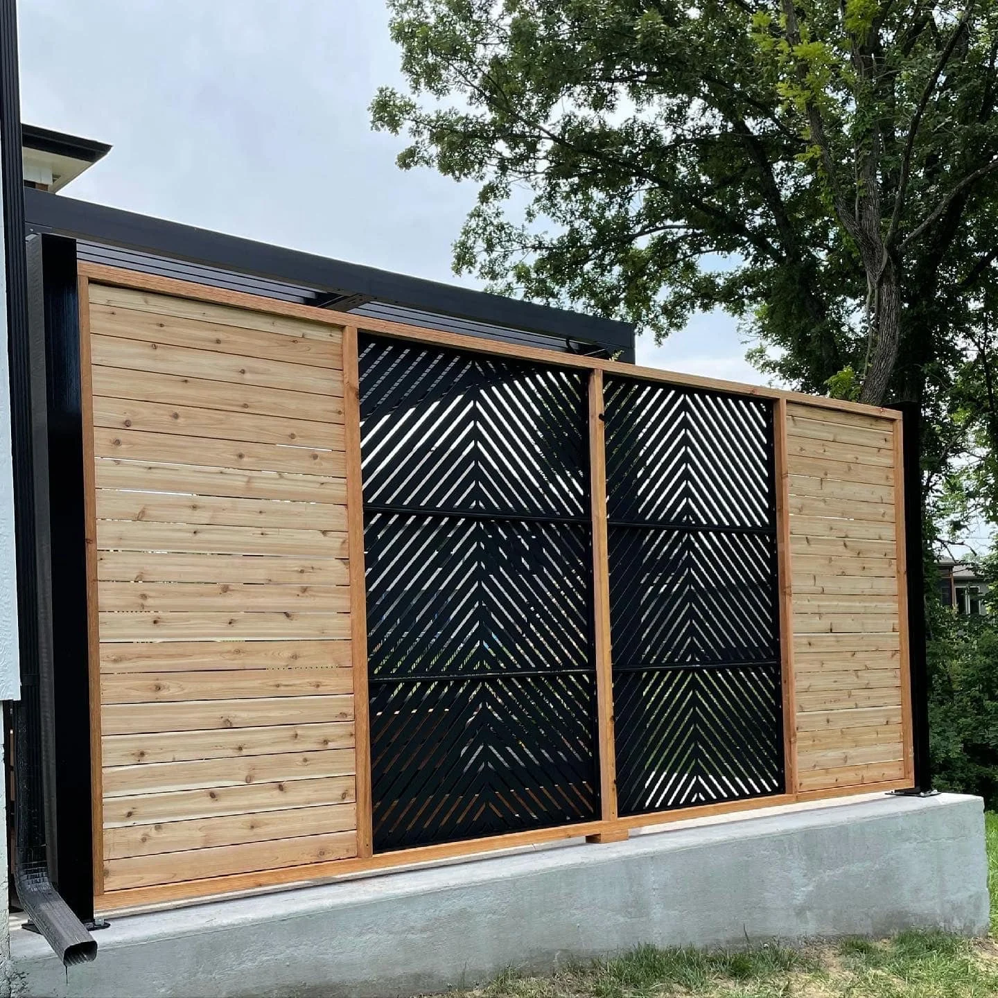 A building with a decorative wooden and black metal fence featuring diagonal slats, set on a concrete base, with a large leafy tree in the background.
