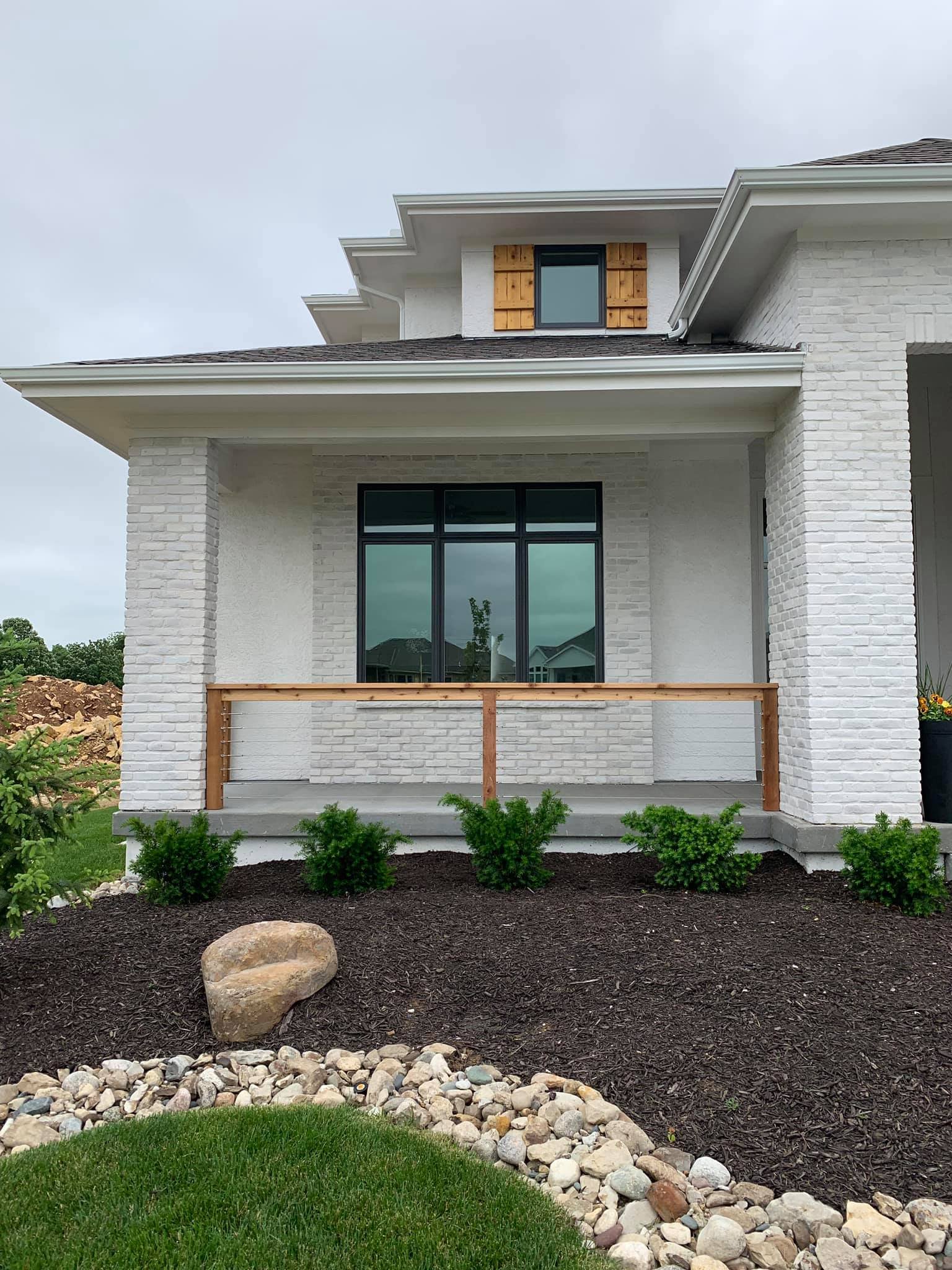 Front view of a white brick house with a small covered porch, large second-floor window, and a smaller window with unfinished wooden shutters. The front yard has mulch, small bushes, a large rock, and decorative stones.