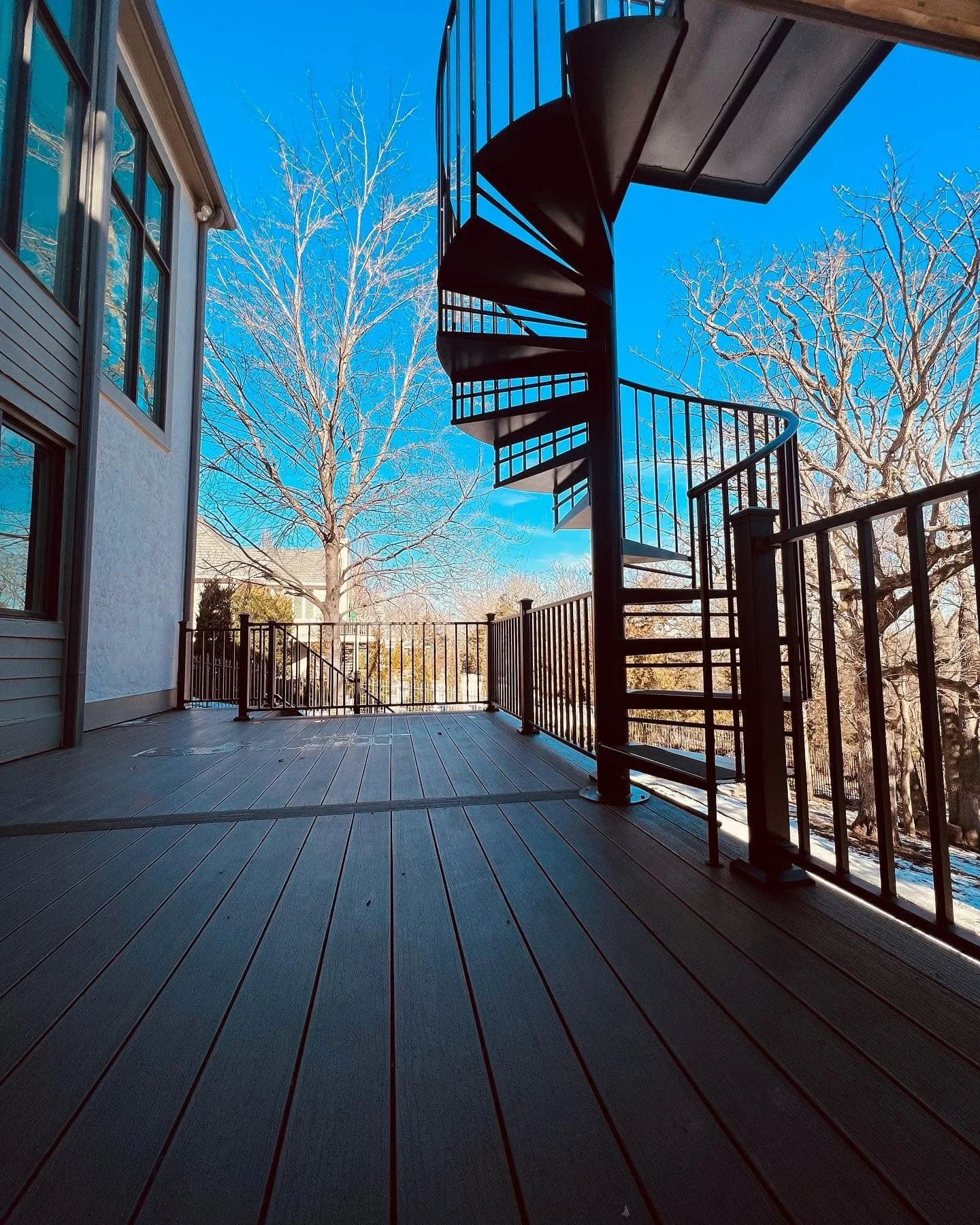 Empty wooden deck with a black spiral staircase on the right and leafless trees in the background, under a clear blue sky.