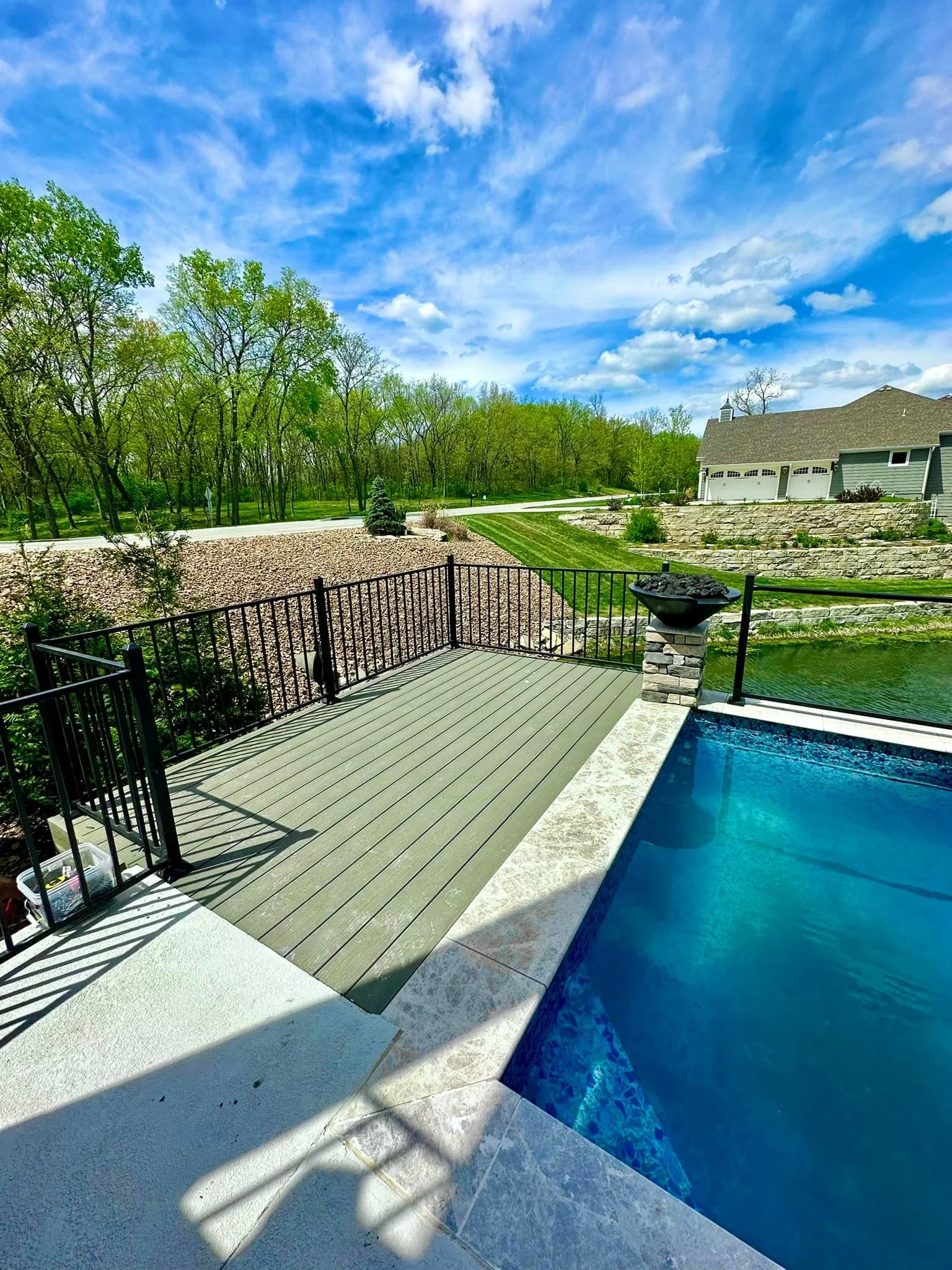 View of a backyard with a swimming pool, a wooden deck, and a black metal fence, overlooking a landscaped yard with trees and a blue sky with clouds.