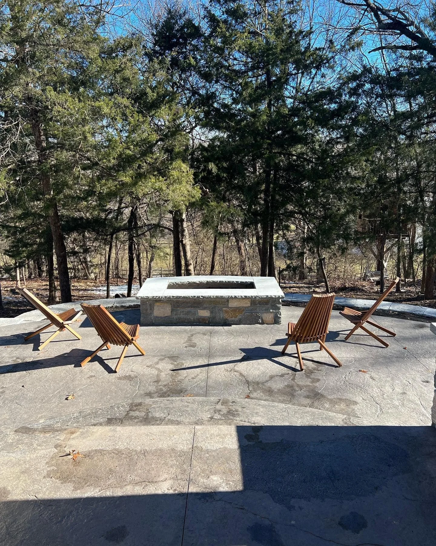 Outdoor patio with five wooden chairs facing a stone fire pit, surrounded by tall trees on a clear day.
