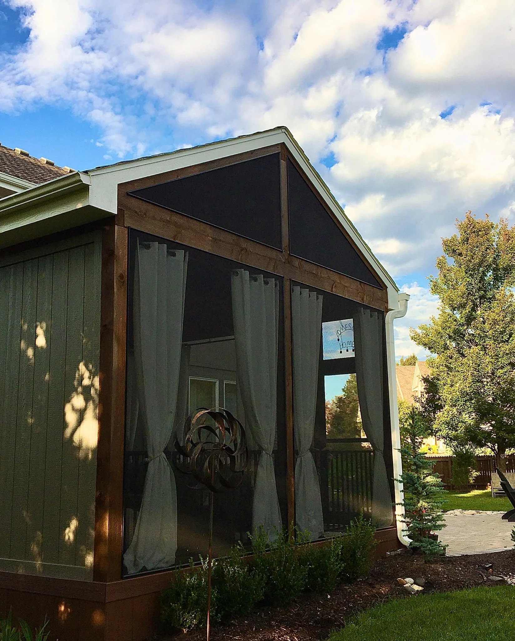 A screened-in porch attached to a house with white curtains, a decorative wind spinner, shrubs, and a small tree in a backyard under a partly cloudy sky.