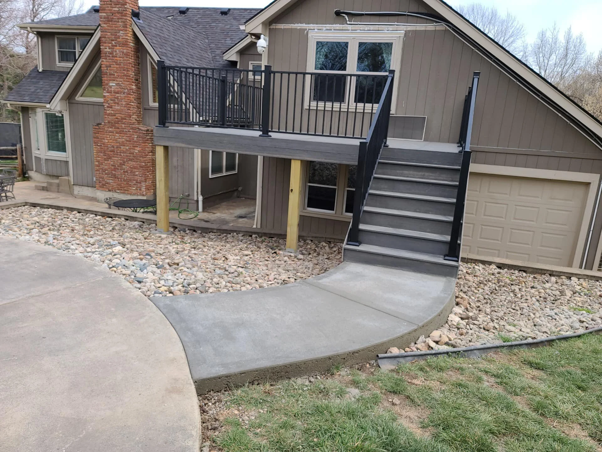 Newly constructed elevated wooden deck with black metal railing and stairs attached to the back of a house. The house has gray siding and a brick chimney. The ground below the deck is covered with rocks and a concrete pathway leads up to the stairs.