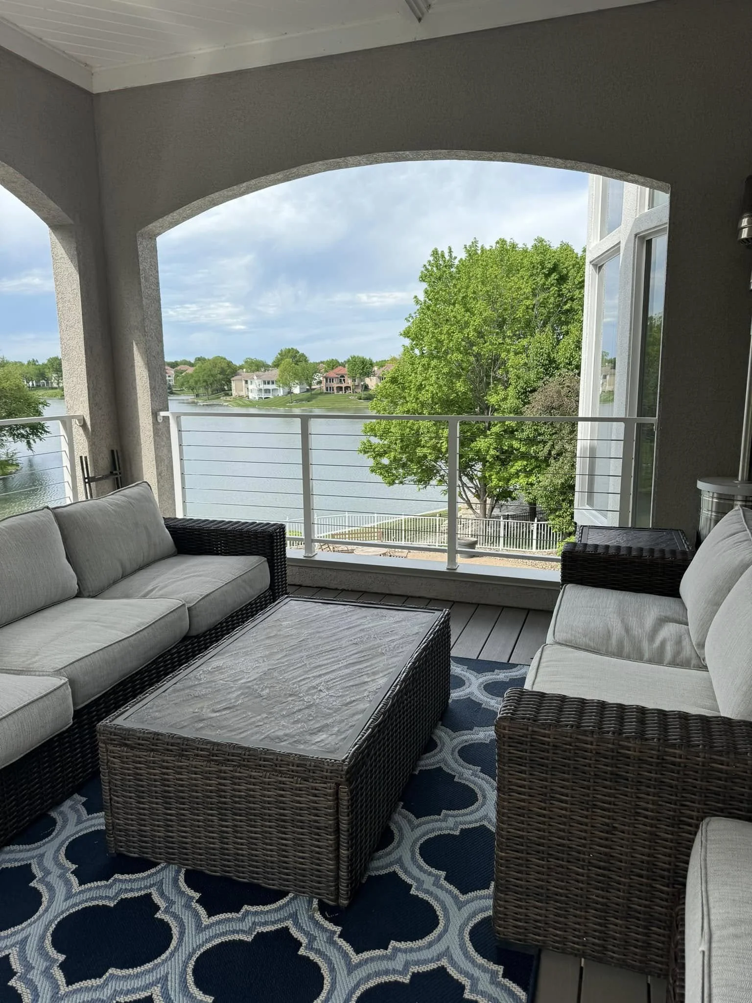 Outdoor balcony with wicker sofa and chairs, a coffee table, and a view of a lake with houses and trees.
