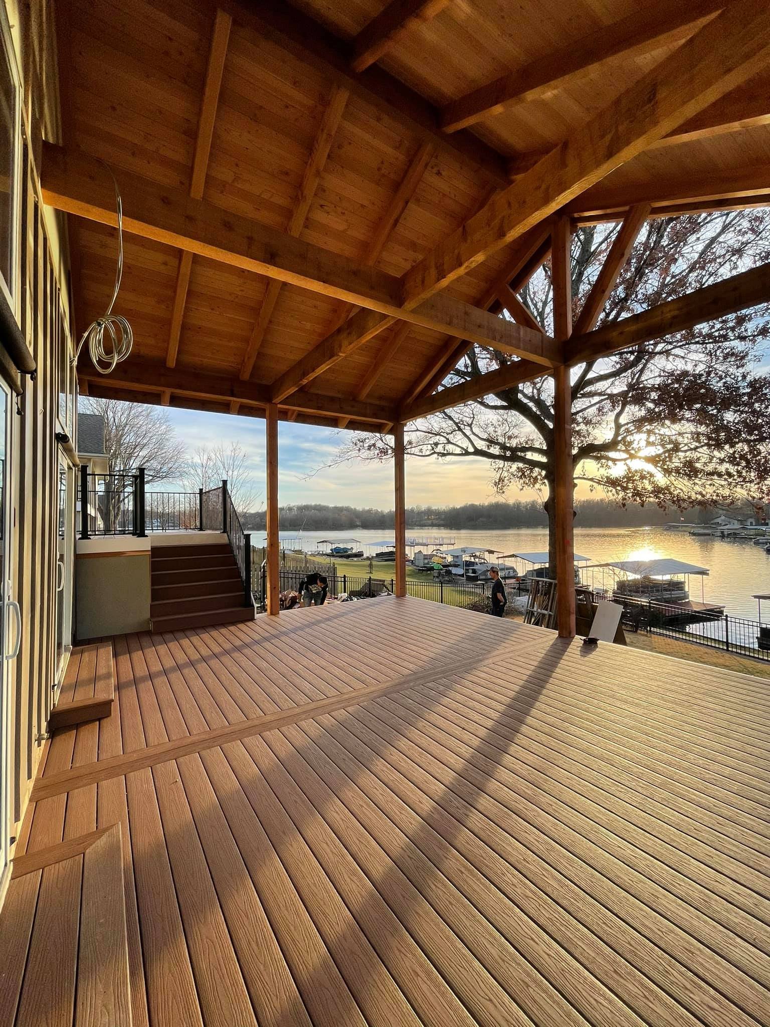 View of a wooden deck with a roof, overlooking a lake with boats and dock coverings, during sunset.