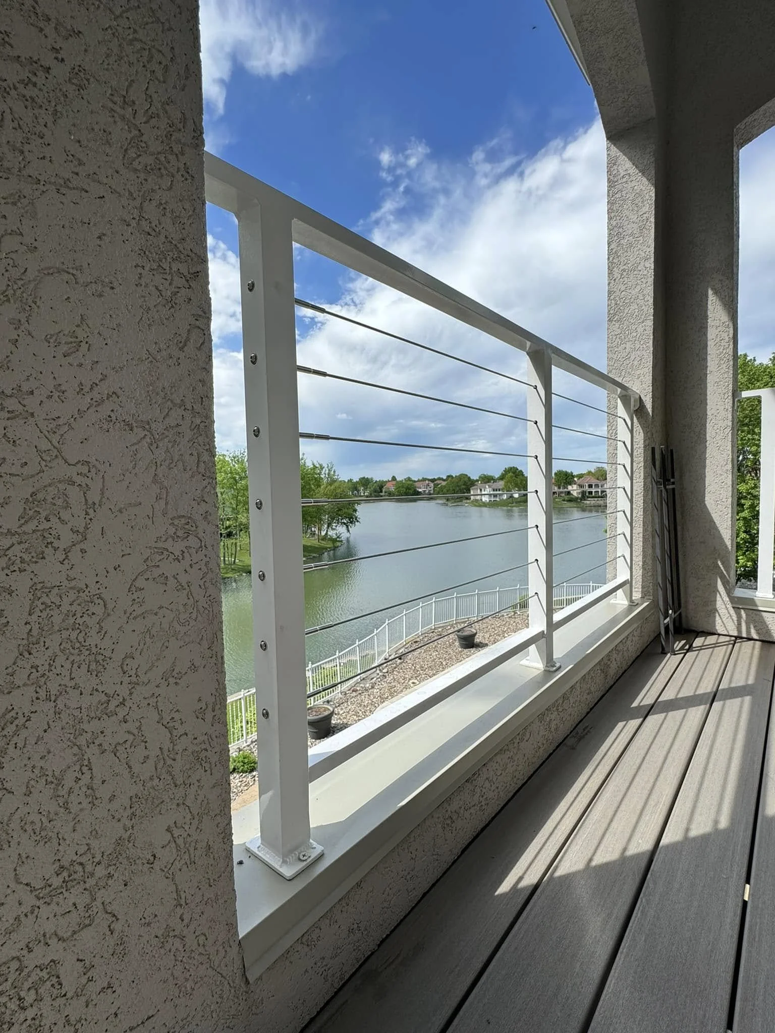 View of a lake from a balcony with a white metal railing, gravel shoreline, and residential houses in the distance under a blue sky with scattered clouds.