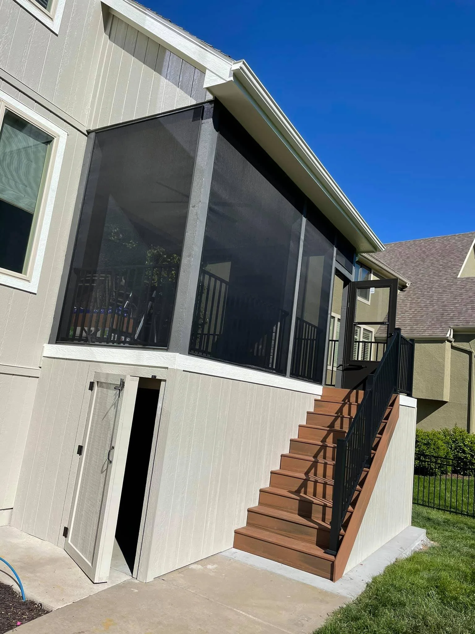 Back porch of a house with wooden stairs, black metal handrails, and screened enclosure on the upper level under a clear blue sky.