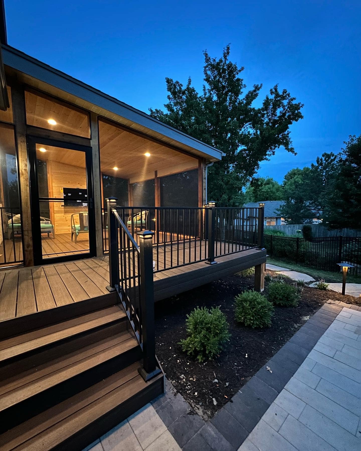 A backyard porch with wooden decking and black railing, illuminated by recessed ceiling lights, with steps leading down to a landscaped area with small bushes and pathway lights, during dusk.