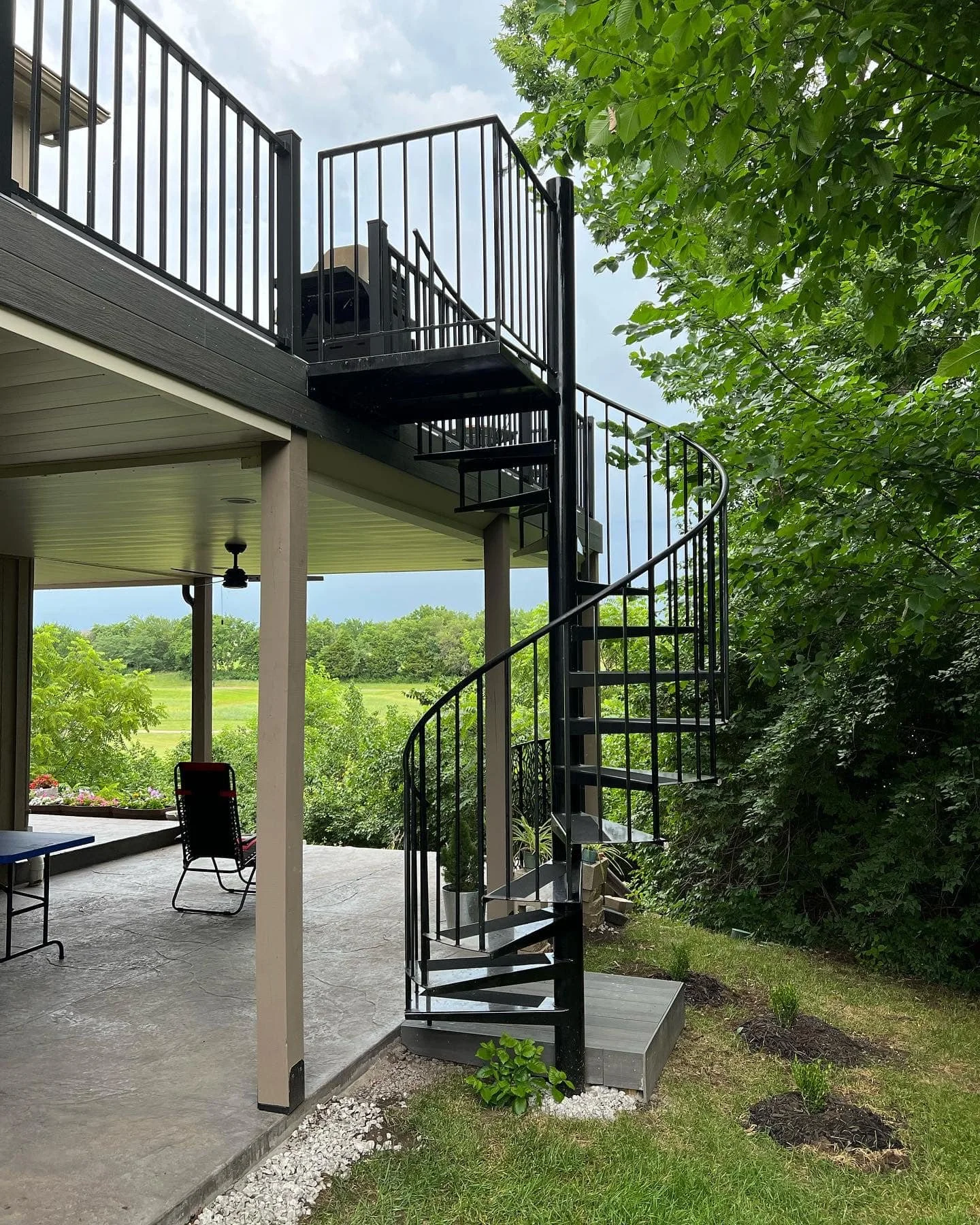 Black outdoor spiral staircase attached to a two-story house with a covered patio, surrounded by green trees and a grassy field in the background.