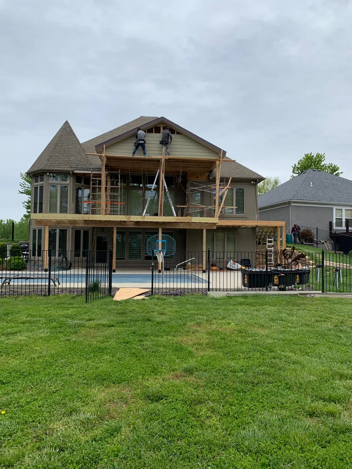 Two construction workers on a wooden balcony of a large house under renovation, with scaffoldings and tools around, in a residential neighborhood with a grassy yard.