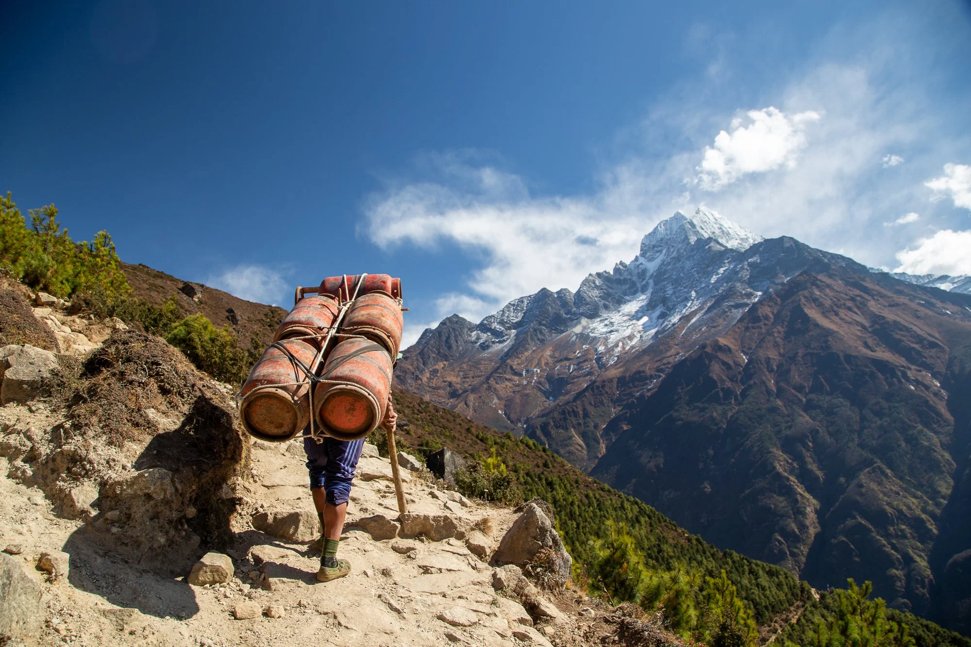 A man hiking on a dirt trail in the mountains, carrying a load of two large, rusty metal fuel containers on his back, with snow-capped peaks and a blue sky in the background.